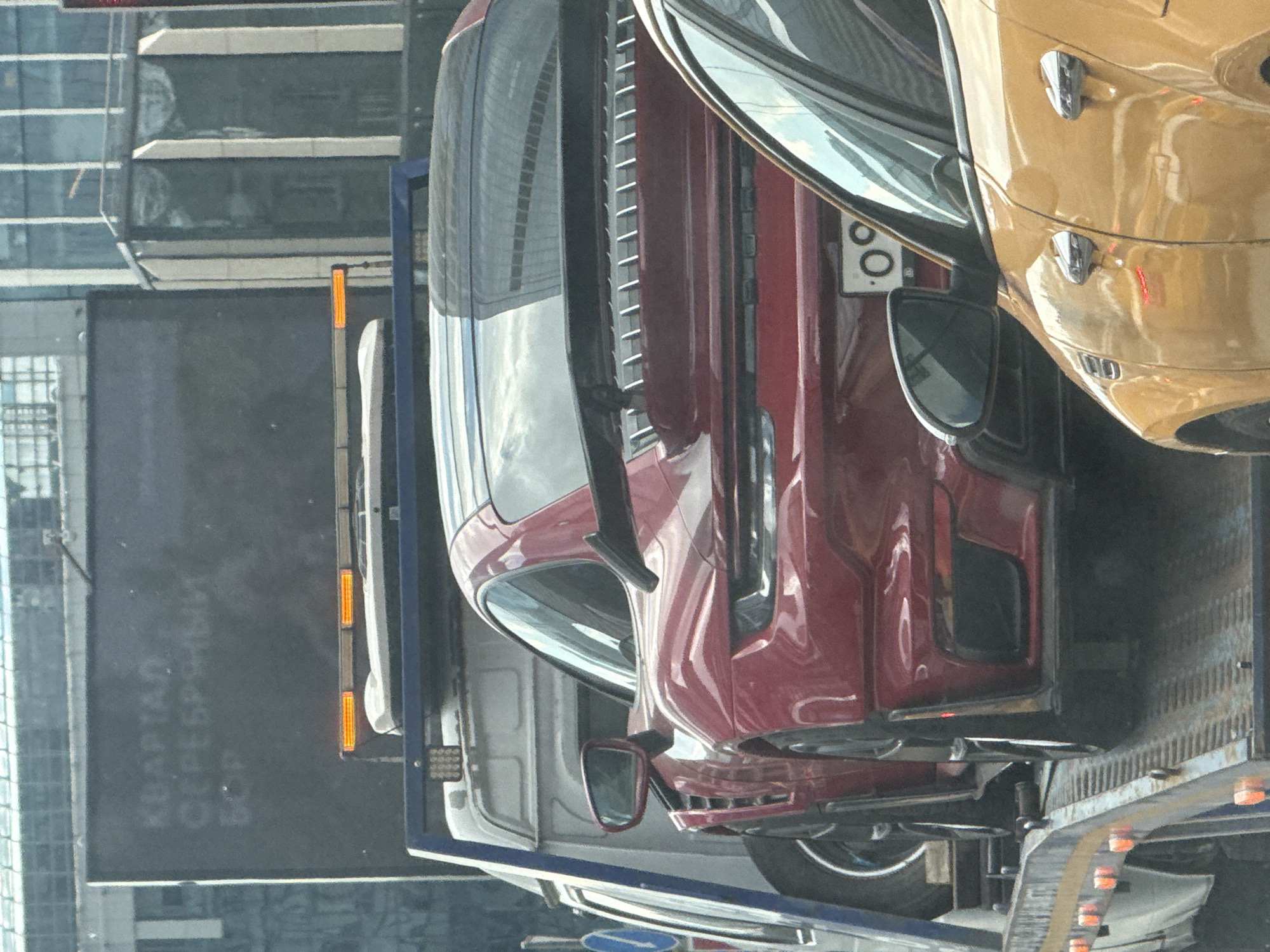 This image, captured in Khimki, Russia, shows a multi-level car carrier truck transporting several vehicles in an urban environment. The perspective is from an elevated viewpoint, looking down onto the truck and its cargo.
The primary subject is a dark red or maroon luxury sedan or coupe, possibly a Cadillac, occupying the lower deck of the car carrier. Its sleek design, prominent front grille details, and side mirrors are clearly visible. Above and slightly to the right of the red car, a gold or tan sports car, exhibiting characteristics similar to a Porsche with its distinctive headlights and door handles, is loaded onto the upper deck. A fragment of a Russian license plate, displaying "00", is visible on the gold car. A white or light-colored vehicle, possibly an SUV or van, is partially visible behind the red car on the lower level.
The car carrier trailer features ramps and orange side marker lights along its structure. The setting appears to be a modern urban landscape during the daytime, with clear weather conditions reflected in the glass facades of tall buildings in the background.
A notable detail is the Cyrillic text "КВАРТАЛ СЕРЕБРЯНЫЙ БОР" (Kvartal Serebryanyy Bor), meaning "Silver Forest Quarter," which is painted in large white letters on the dark asphalt in the foreground, providing a specific location reference within Russia. The image captures the activity of professional vehicle transport in a city setting.