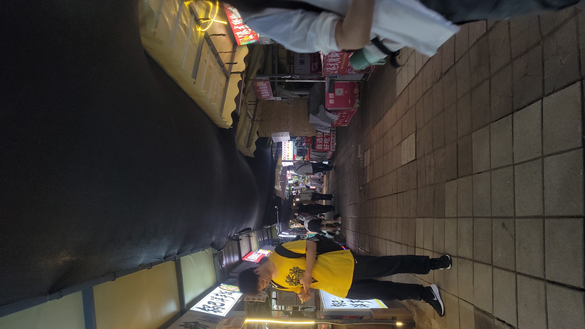 The image captures a bustling narrow street or market at night, illuminated by artificial lights from numerous stalls. Several people are visible, engaged in activities typical of a market setting.
In the foreground, a person wearing a bright yellow t-shirt with a dark graphic and black pants stands on the left side of the frame, appearing to look at one of the stalls. This individual is wearing glasses. The ground is paved with square-tiled or cobblestone-like material.
On both sides of the path, various stalls or small shops are set up under awnings, displaying items and signs. The lighting indicates it is evening or night, creating a lively atmosphere. People are seen further down the path, walking or congregating near the stalls. The overall setting suggests an urban environment, likely in East Asia due to the presence of Chinese script.
Visible text on signs includes: "POCC0000", "自选快" (self-selection quick), "好吃下" (delicious), "9.9半斤,米饭一元" (9.9 half jin, rice one yuan – indicating pricing for food), "重庆" (Chongqing, likely referring to cuisine), "泰奶" (Thai milk, likely Thai milk tea), and "港式猪脚" (Hong Kong style pork trotters). These texts suggest a variety of food options available in the market.