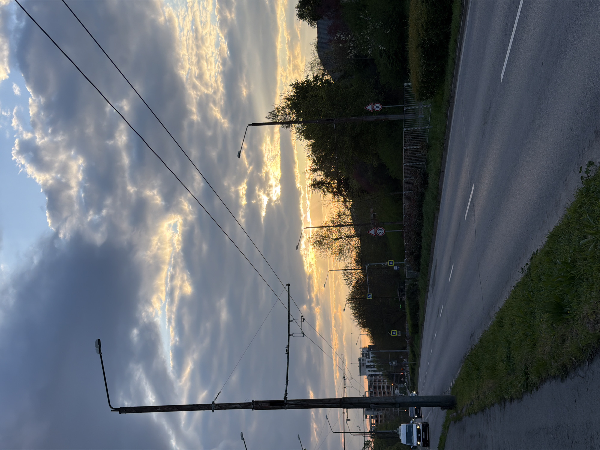 The image depicts an outdoor urban scene at either sunrise or sunset, likely in Varna, Bulgaria. The sky is a prominent feature, filled with dramatic, textured clouds. Warm golden light from the sun, positioned towards the right side of the frame, breaks through these clouds, illuminating their edges and creating striking contrast with the darker, cooler blue-grey clouds on the left.
In the foreground and mid-ground, a network of utility poles and overhead electrical lines, characteristic of trolleybus or tram infrastructure, extends across the upper and central parts of the frame. Streetlights are mounted on several of these poles.
To the left, an asphalt road with white dashed and solid lane markings stretches into the distance. A narrow strip of green grass borders the immediate left edge of the road. Further left, an embankment covered with dense trees and bushes rises. Along this embankment, several traffic signs are visible: multiple circular signs with a red border and the number "30" (indicating a 30 km/h speed limit), and yellow square signs with a black pedestrian symbol (pedestrian crossing). A metal fence or barrier also runs alongside the road.
In the bottom-left corner of the road, a white van, possibly a Ford Transit, is partially visible, facing towards the viewer. Another vehicle can be seen further down the road. In the far distance, beyond the treeline, glimpses of urban buildings are visible. The overall mood is serene, capturing the unique light of dawn or dusk over a city street.