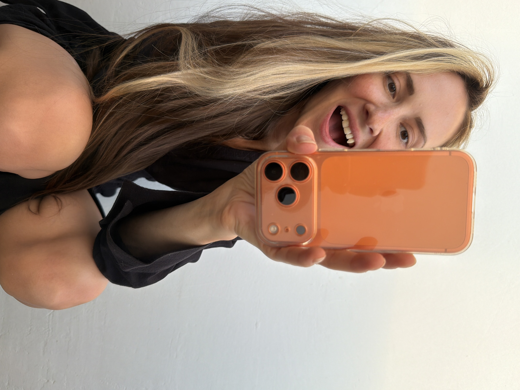 A woman is taking a selfie with an orange smartphone. She is crouched, holding the phone out at arm's length. Her hair is long and blonde, with brown underneath. She is wearing a dark-colored top. The background is a plain white wall. There is no evidence to suggest this photo was taken in Khimki, Russia.