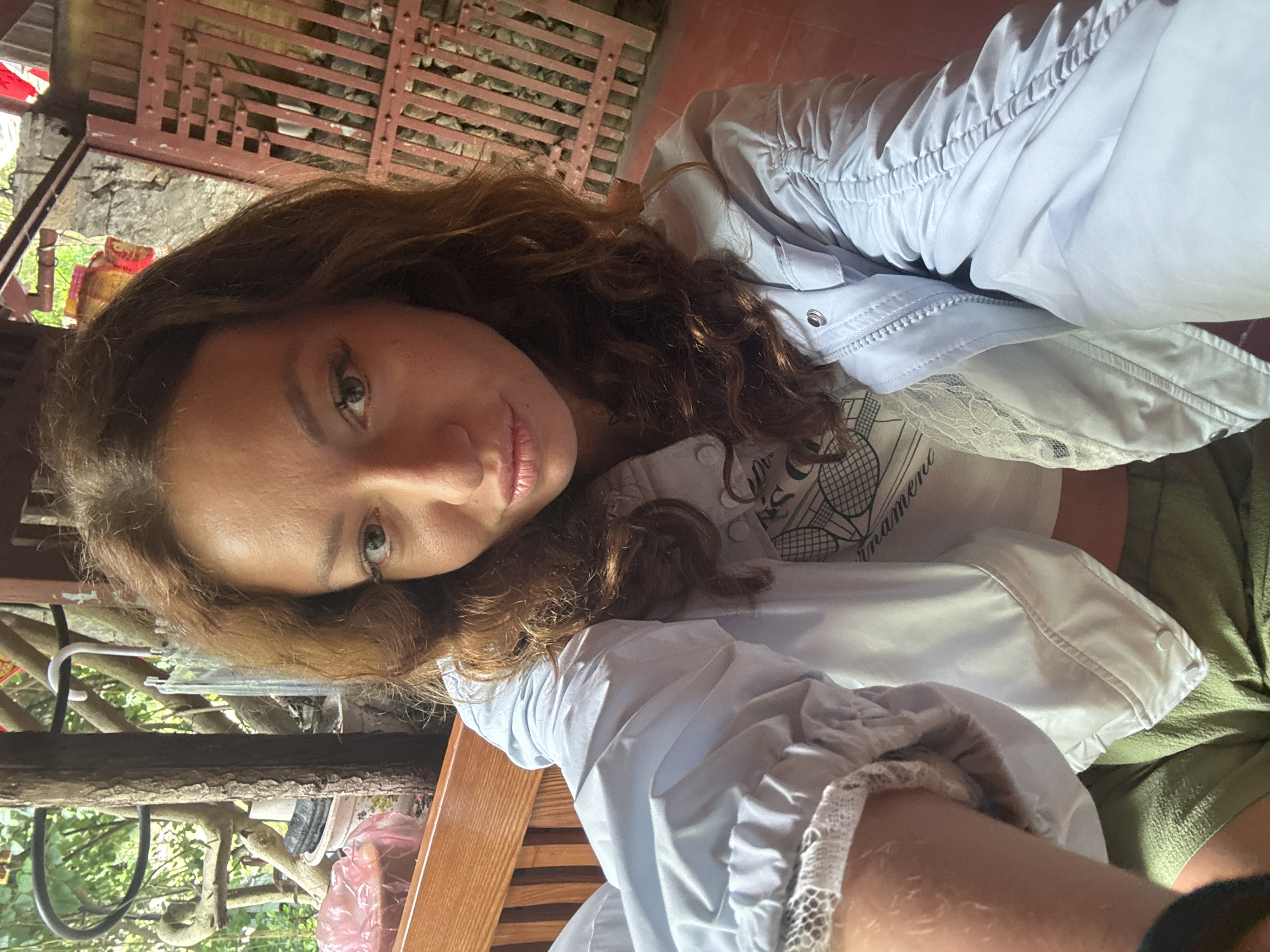 A woman poses indoors in Mèo Vạc Commune, Vietnam. She has brown curly hair and light skin. She wears a light blue jacket over a white t-shirt and green bottoms. The setting includes a rustic wooden structure, likely a building, with a reddish-brown lattice wall in the background. Other wooden elements, possibly a fence or structure, are also visible with glimpses of greenery. The lighting is soft and natural.