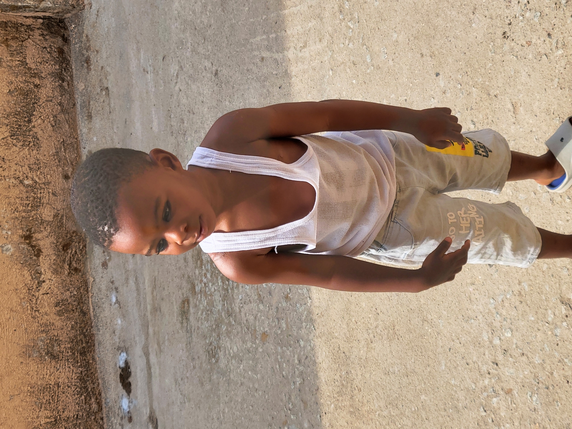 A young, dark-skinned boy with short, dark hair stands on a concrete surface. He is wearing a white tank top and light gray shorts with faded writing on the right leg. He appears to be standing on a street or sidewalk, with a textured, tan concrete surface behind him and a darker, possibly building wall, along the left edge of the image. One white and blue sandal is visible on his right foot. It is daytime. This scene is located in the city of Suleja, Nigeria.