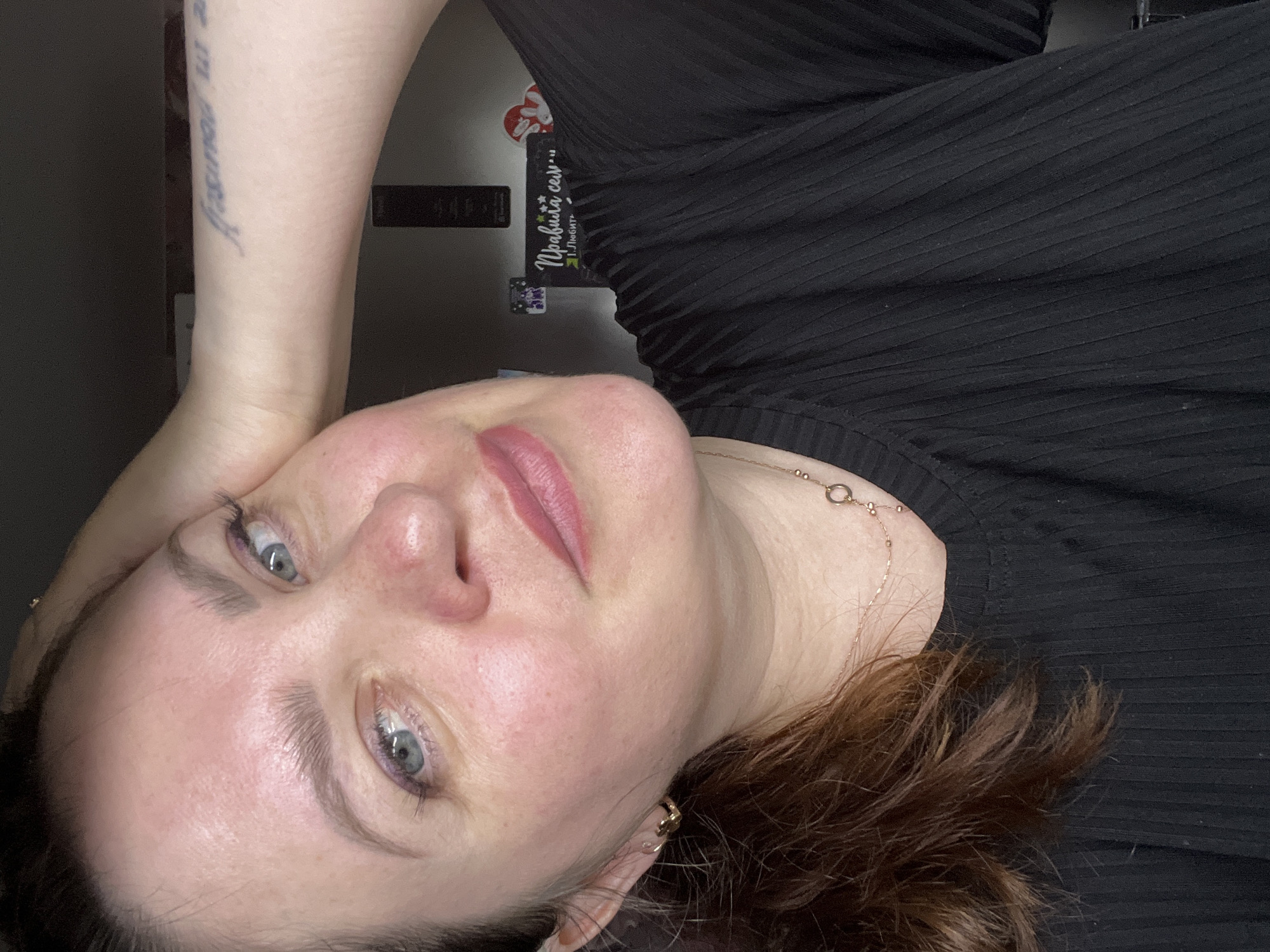 A female subject, appearing to be an adult, is depicted in a close-up, chest-up self-portrait. She has fair skin with some visible redness on her cheeks and nose, blue/grey eyes, and dark brown hair that appears pulled back, with some loose strands framing her face. Her eyebrows are well-defined, and she has light purple eyeliner or eyeshadow on her lower lash lines. Her lips are a natural pink color, showing a slight smile. She wears a black, ribbed, short-sleeved top and accessorizes with a delicate gold-colored chain necklace featuring small beads and a circular pendant, as well as a small gold hoop earring in her visible left ear. Her right arm is raised, with her hand resting on the back of her head, revealing a tattoo in a script font on her right forearm that appears to read "Believe in yourself" (partially visible).
The background suggests an indoor setting, characterized by a dark, indeterminate surface. On the right side of the frame, behind the subject's shoulder, a black sign or poster is visible with white Cyrillic text. The legible text includes "Правила семьи" (Rules of the family) and below it "1. Любите" (1. Love). Several small stickers are also affixed to this surface.
The specific city of Noviy, Russia, is not discernible from the image content. The environment is a domestic or personal space.