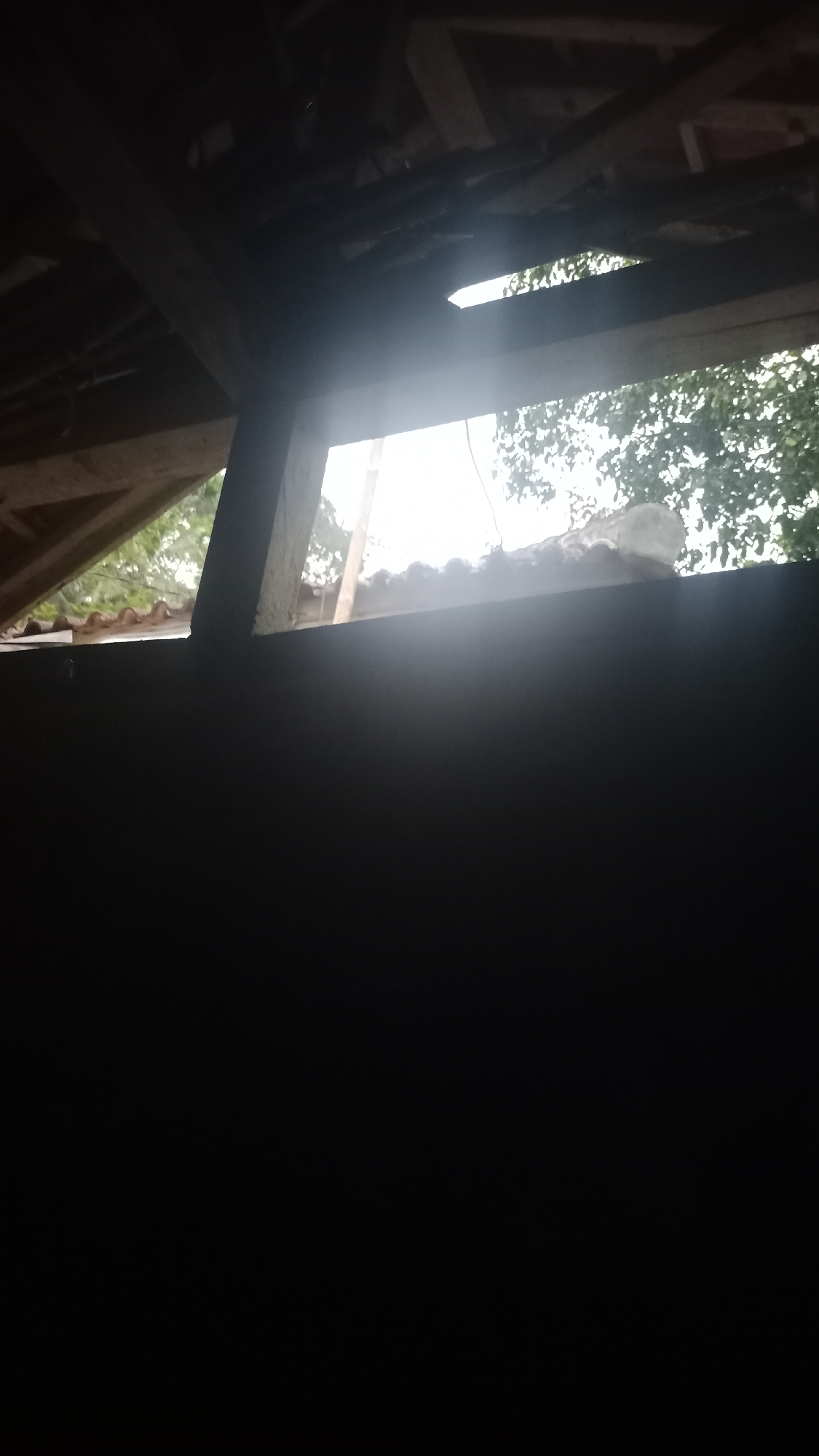 The image shows a partially constructed roof structure with exposed wooden beams and roof tiles. Sunlight streams through gaps in the roof, illuminating the interior. The lower portion of the image is dark, suggesting it is inside a building or under an overhang. The background shows foliage and the sky, indicating an outdoor setting. The image appears to be taken in Desa Burujul Jaya, Indonesia.