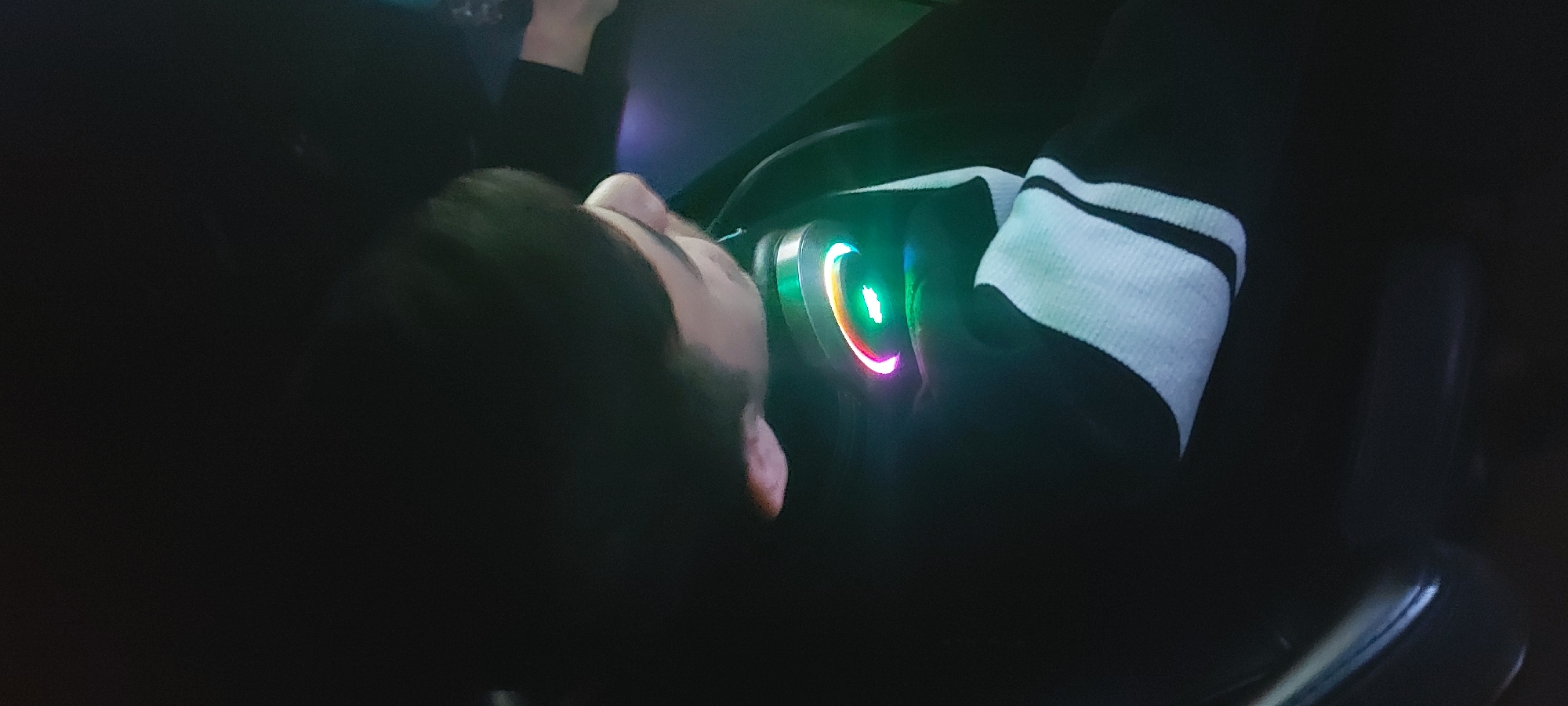 The image appears to be taken indoors in a dimly lit environment. A person is visible, with their face and hair partially illuminated. They are wearing headphones with rainbow colored lights. The person's arm, covered by a black sleeve and a white sleeve with black stripes, is positioned near the headphones. A chair is partially visible in the lower right corner of the image. It is not possible to verify or comment on the location of this image in the city of Tashkent, Uzbekistan, based on the provided visual information.