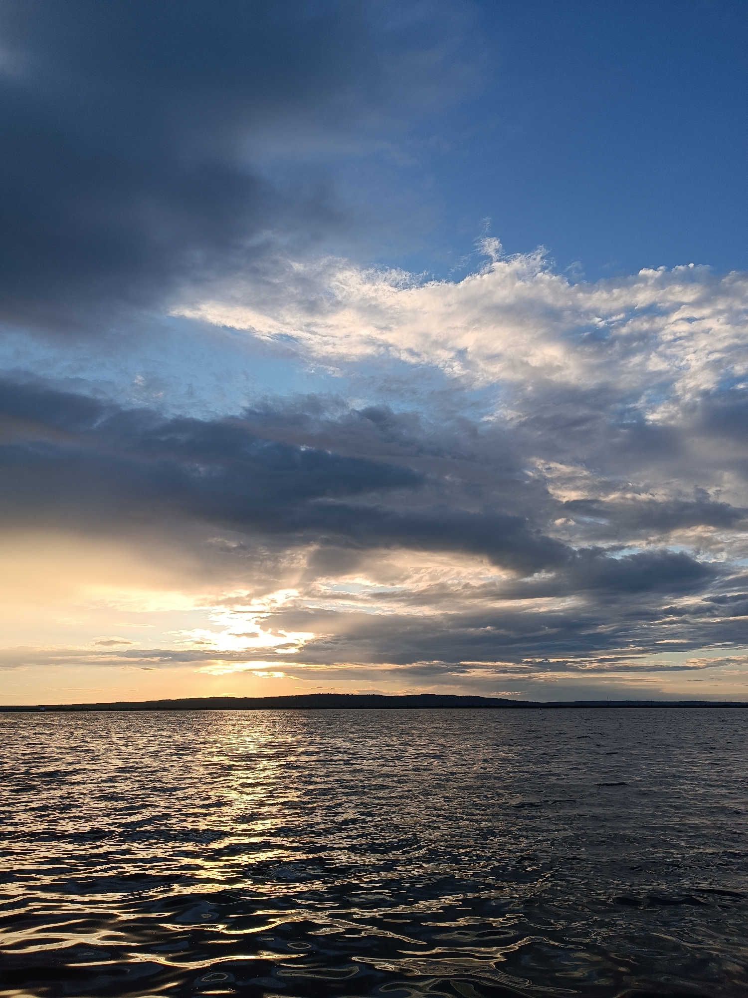 A serene, vertical landscape captures the transition from day to evening over a body of water. The lower portion of the image is dominated by the dark, rippling surface of a lake or sea, reflecting the light from the sky. The water's surface is textured with small waves, creating a shimmering effect where sunlight catches the crests.
The horizon line is low in the frame, separating the water from the sky. A distant landmass, possibly a shore or rolling hills, is visible as a dark silhouette against the brighter sky.
The sky is a dramatic canvas of clouds and fading sunlight. The upper part of the sky is a clear, deep blue, transitioning into patches of lighter blue and scattered white clouds. Towards the horizon, the clouds become more dense and varied in color, ranging from dark grays and blues to softer whites and yellows. Sunlight breaks through in several places, casting rays and illuminating portions of the clouds from behind. The overall impression is of a beautiful, dynamic sunset or sunrise.
The time of day appears to be either early morning or late evening, given the soft, angled light. The weather seems to be changing, with evidence of both fair and possibly stormy weather in the cloud formations. The location cues of Chaikovsky, Russia, suggest a natural, possibly rural or semi-urban waterfront setting. The emotion evoked is one of peace, awe, and the natural beauty of the environment. There is no visible text in the image.