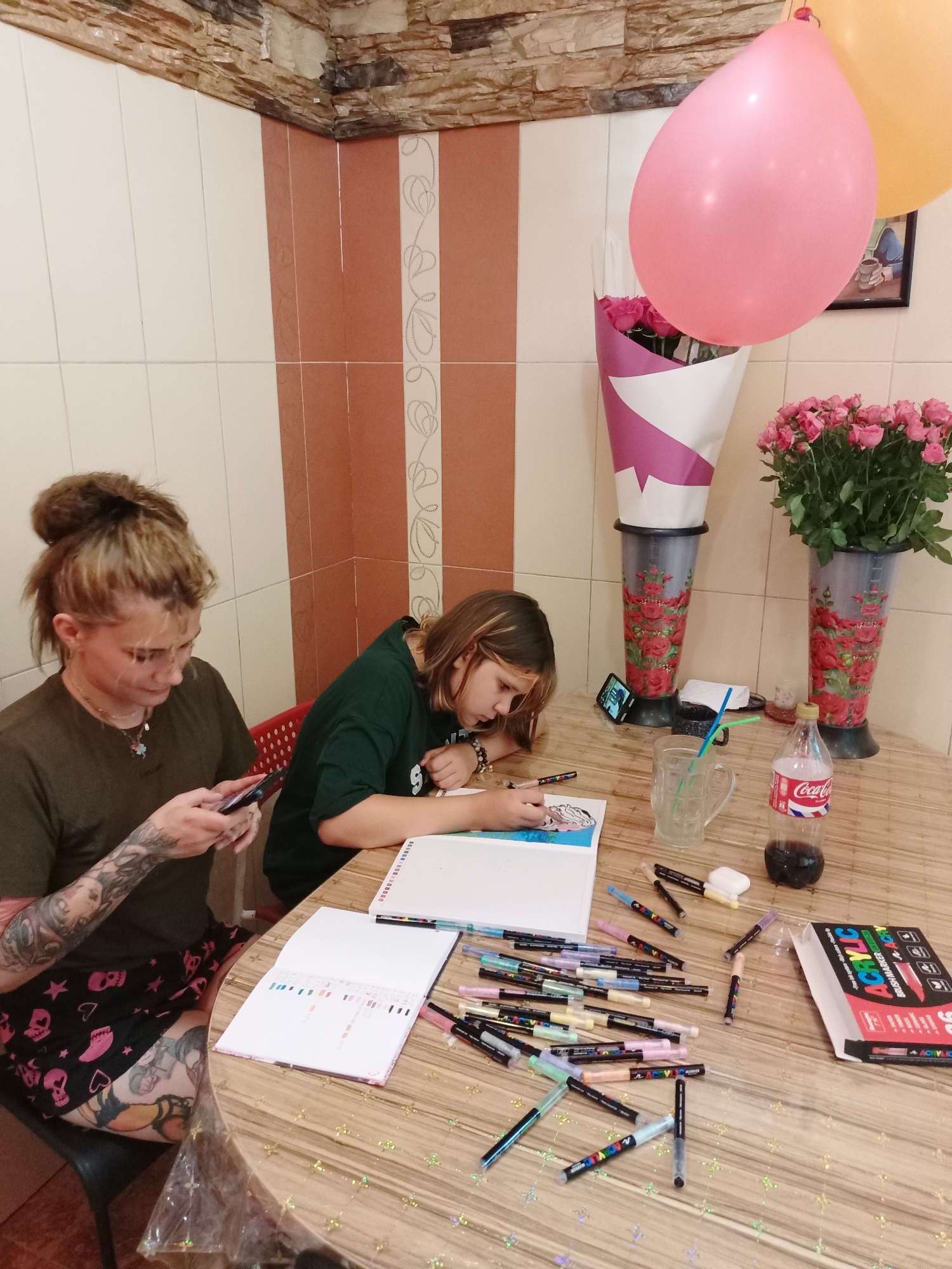 The image depicts an indoor scene, possibly a home or a small creative space, with two individuals engaged in artistic activities at a round wooden table. The room features tiled walls, with a section in the background showing a mix of white and reddish-brown tiles, and a decorative element with a floral pattern. The ceiling has a rustic, stone-like texture.
On the left, a person with blonde hair pulled up in a bun, wearing a dark green t-shirt, is looking down at a smartphone in their hands. This individual has numerous tattoos on their arms and legs, visible on their left arm and leg. They are seated on a red plastic chair.
To the right, a younger person with light brown hair is focused on drawing in a sketchbook placed on the table. They are wearing a dark green t-shirt with a white "S" logo on the back. Their right arm is resting on the sketchbook as they draw.
The table is covered with art supplies, including a large collection of colorful marker pens scattered around. A sketchbook with color swatches is open in front of the second person. Also on the table are a glass of liquid with straws, a bottle of Coca-Cola, and a box labeled "ACRYLIC BRUSHMARKER." In the background, two vases hold bouquets of pink roses. Two balloons, one pink and one yellow, are suspended in the upper right portion of the image. The overall impression is of a creative session, possibly a birthday celebration given the balloons and flowers.
The image does not contain any visual cues that would allow for the identification of the city of Almaty, Kazakhstan.