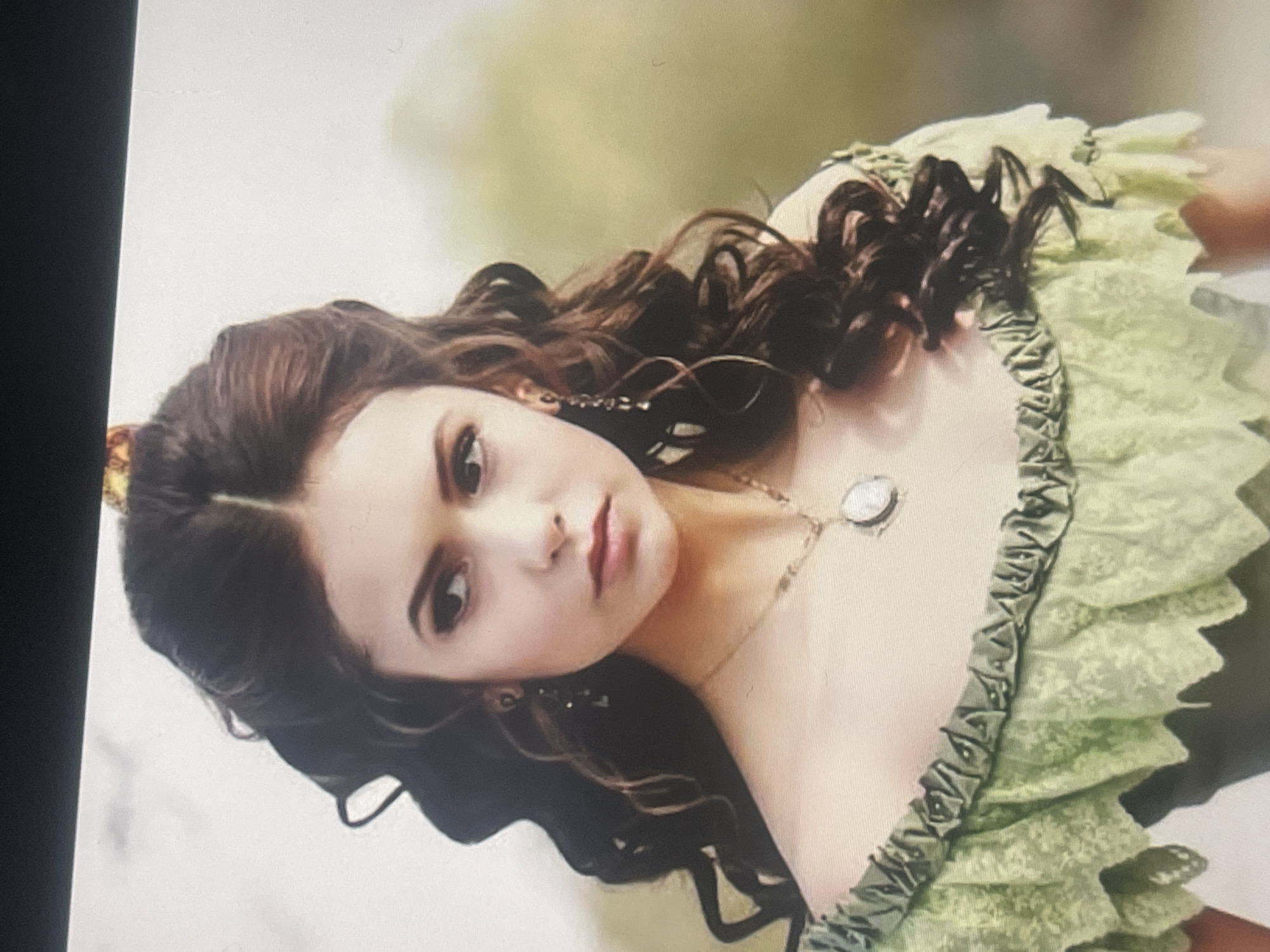 This image features a young woman posing outdoors, with the provided context indicating the location is Poltava, Ukraine. She has long, dark brown, curly hair, parted down the middle, and fair skin. Her face is made up with prominent eye makeup and dark reddish lipstick. She is wearing a light green, off-the-shoulder garment, possibly a dress or top, detailed with ruffled or laced layers along the neckline and sleeves. A thin golden necklace with a round, locket-style pendant adorns her neck, and she wears long, dark earrings.
The woman gazes directly at the viewer with a serious or slightly pensive expression, her head tilted to one side. The background is softly blurred, showing shades of green and lighter hues, indicative of foliage or a natural outdoor environment during the daytime. A dark vertical bar is visible along the left edge, suggesting the image might be a photograph of a screen or part of a digital display. There is no visible text in the image.