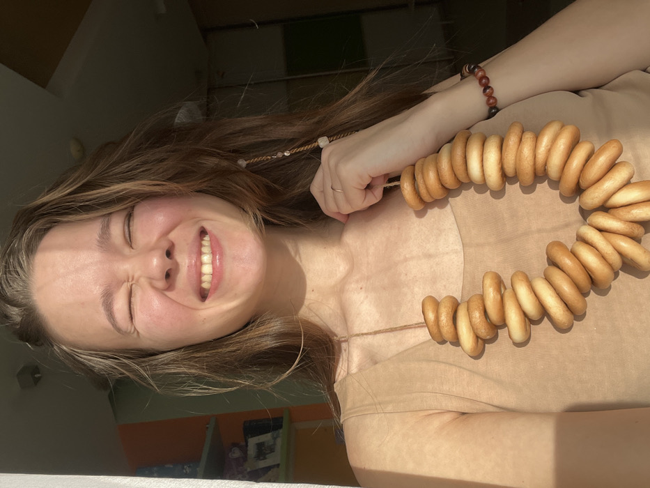 A woman with brown hair smiles, eyes closed, as she wears a necklace of bagels. She is wearing a beige shirt, and her left wrist has a beaded bracelet. Sunlight illuminates her face. In the background, a room with colorful walls and a window is visible. The image is taken from a slightly overhead angle.