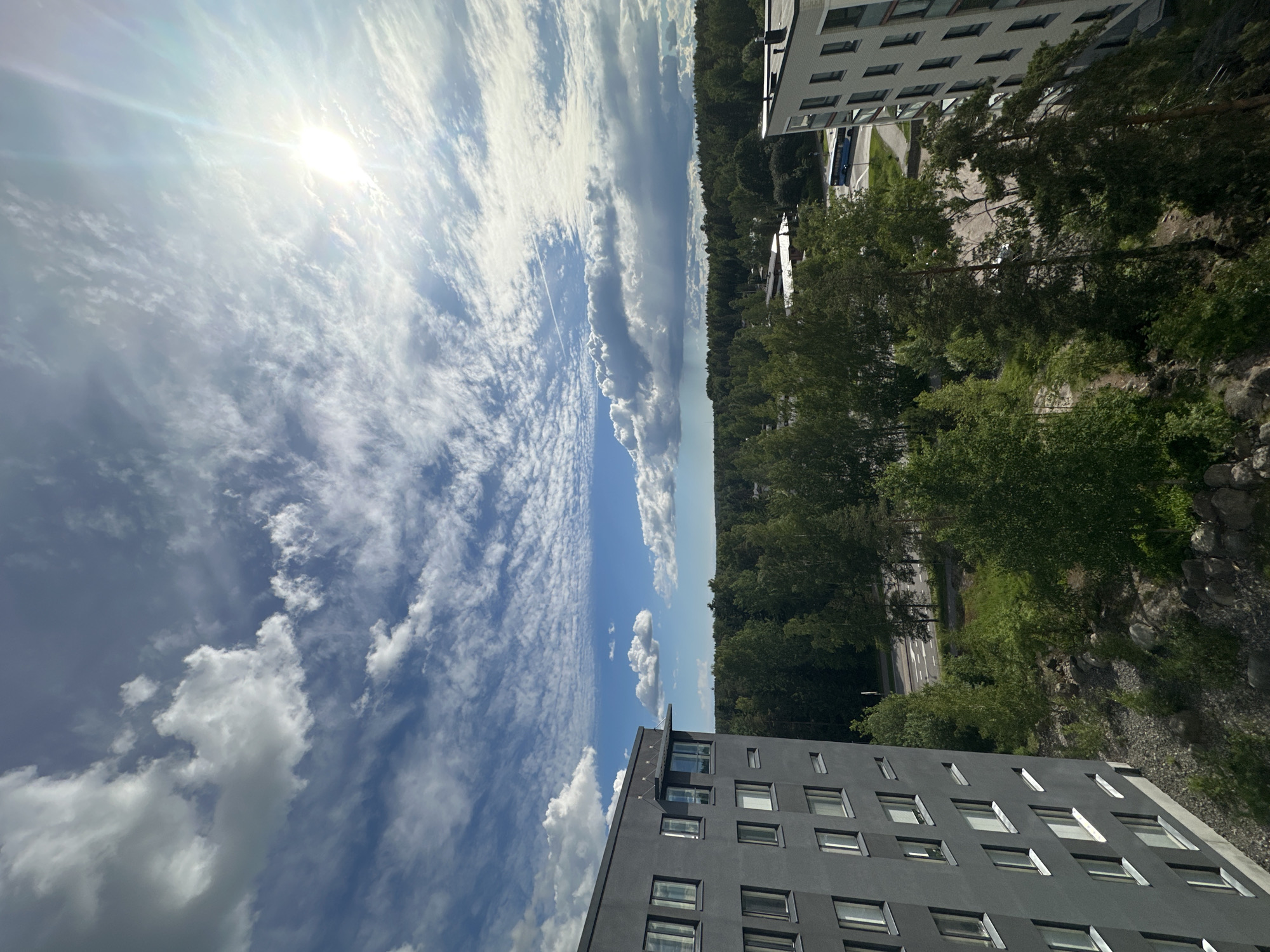 The image is an aerial view of a suburban area with two apartment buildings, a dense forest, and a road. The sky is partly cloudy with the sun visible in the upper right quadrant. The building on the left is a tall, dark gray structure with numerous windows. The building on the right is a lighter colored structure with balconies. The forest is composed of various trees, appearing green and lush. A road with some traffic is visible in the middle ground. The scene depicts an outdoor environment on a bright, partly cloudy day.