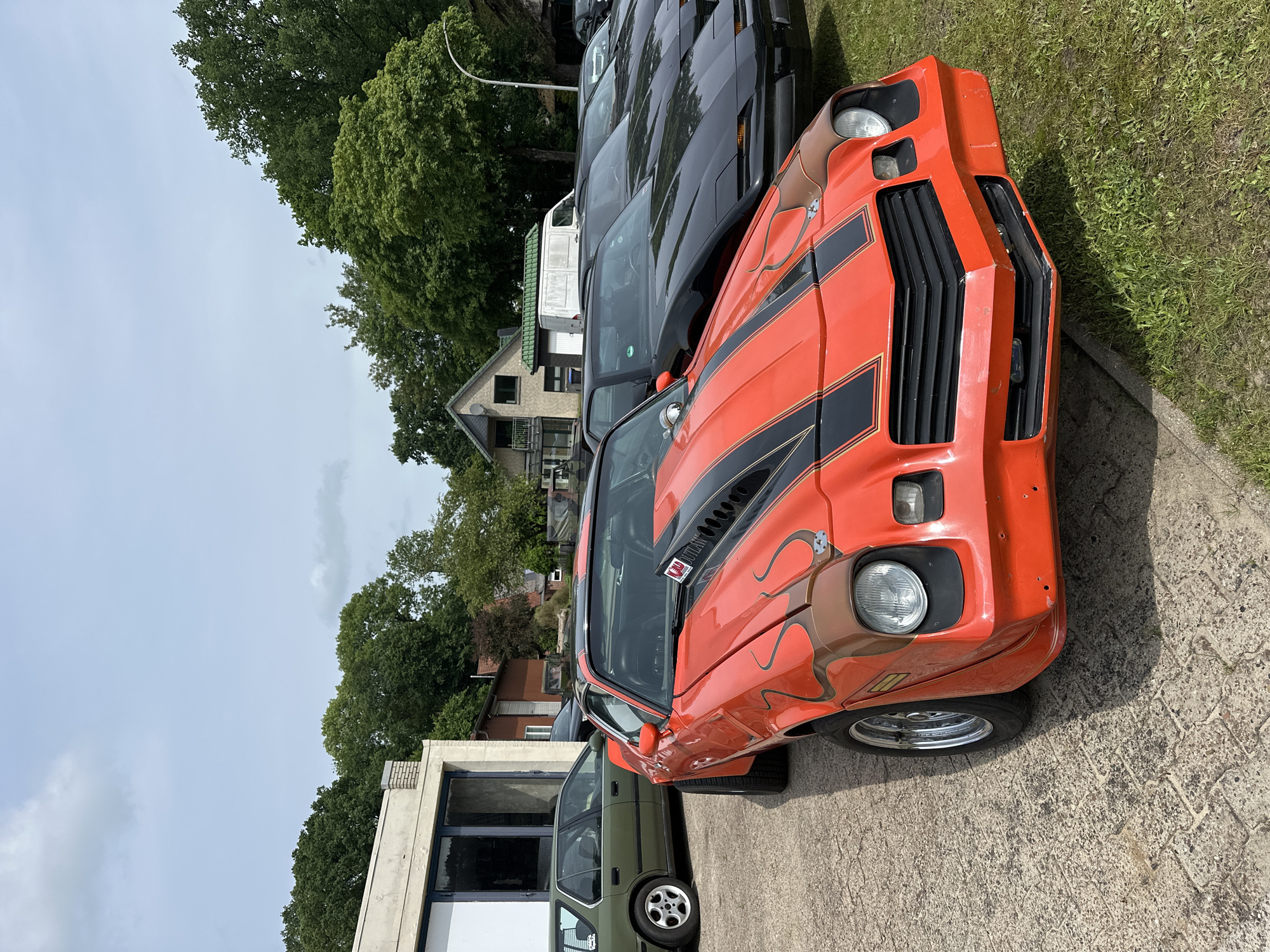 An orange 1970s muscle car with flame graphics and a black racing stripe is parked on a paved area next to a grassy lawn. In the background, a row of black Pontiac Firebirds are parked. Behind the cars, a building with large windows and a white trim can be seen. Green trees and a cloudy sky are visible in the background. The scene appears to be a car show or a gathering of car enthusiasts, possibly at an outdoor venue in Kutenholz, Germany. There are no people visible in the image.
