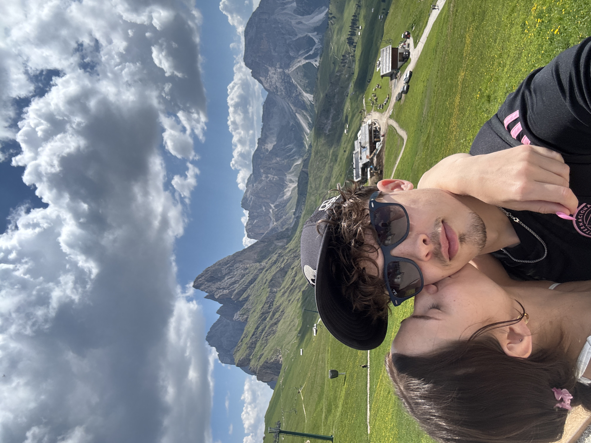 The image features a close-up selfie of a young couple in a mountainous landscape. The man, on the left, wears sunglasses and a baseball cap, and he leans in towards the woman, who is on the right. The woman is resting her head on his arm and appears to be kissing him on the cheek. Both are smiling, conveying a sense of affection and joy.
The background reveals a breathtaking vista of the Dolomites in Santa Cristina Gherdëina, Italy. Lush green meadows dotted with yellow flowers stretch across the slopes, leading up to imposing, jagged mountain peaks. Several buildings, likely chalets or mountain huts, are nestled into the landscape, suggesting a tourist or recreational area. A ski lift is also visible, hinting at the location's dual seasonal appeal. The sky is a brilliant blue, interspersed with dramatic, fluffy white and gray clouds, indicating fair weather with some cloud cover. The lighting suggests it is daytime, possibly late morning or early afternoon, given the brightness and the shadows cast.
On the man's clothing, the word "INTERNACIONAL" is partially visible on his sleeve, along with pink stripes. The woman has a pink hair tie and a small earring. The overall mood is romantic and idyllic, capturing a moment of shared happiness amidst a stunning natural environment.