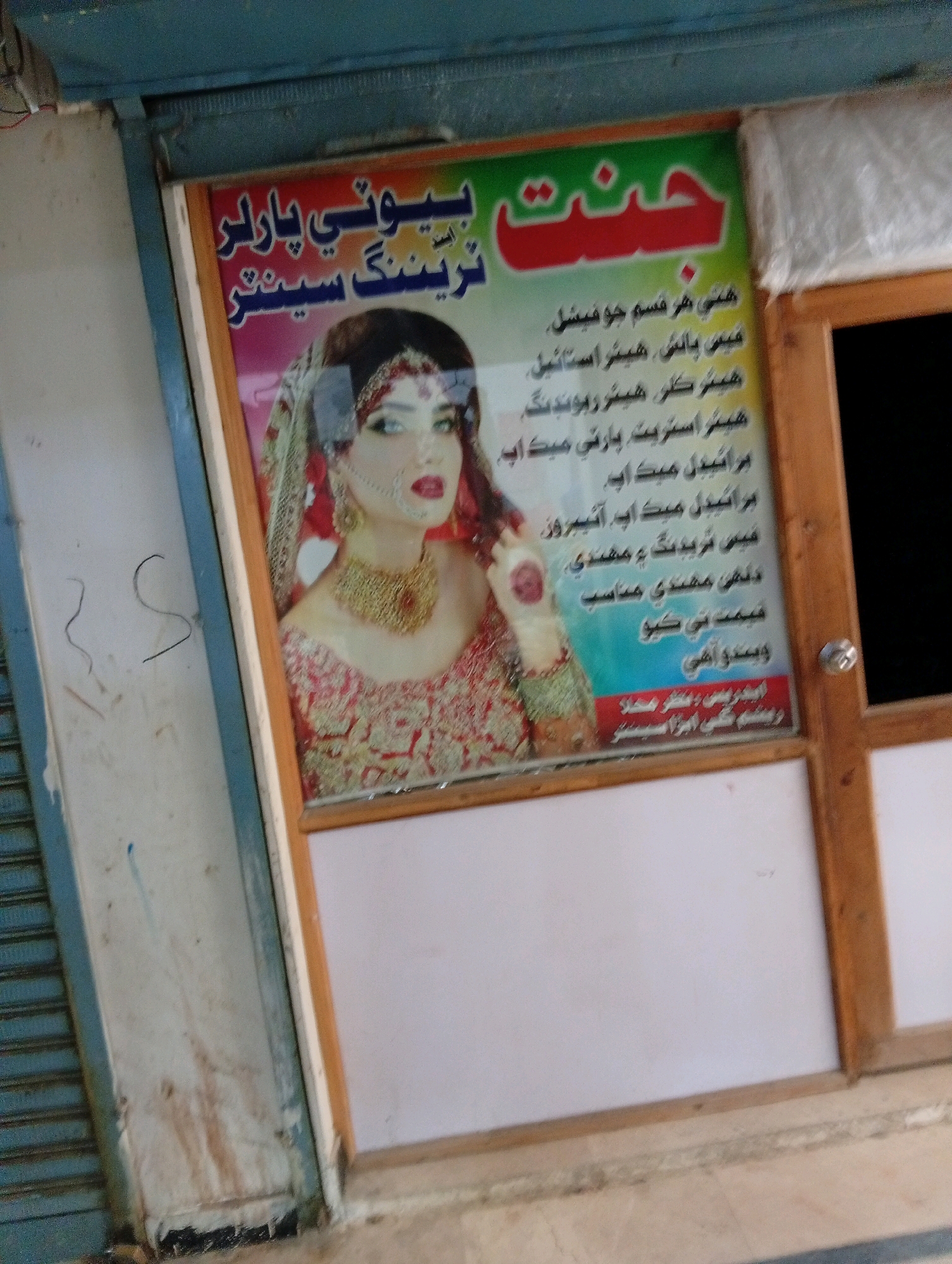 The image shows a storefront in Larkana, Pakistan, featuring a sign for a beauty salon. The sign, primarily in Urdu script, advertises services such as "Gent" beauty parlor, "hair styling," "hair cut," "party make up," "bridal make up," "threading," and "mehndi." A photograph of a woman in bridal attire, with heavy makeup and jewelry, is displayed on the sign. The storefront itself appears to be a modest establishment, possibly in a commercial area of the city. The building has a blue metal shutter, a wooden frame around the sign, and a white panel underneath. The overall impression is that of a typical local business in Pakistan.