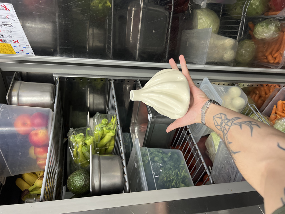 The scene depicts the interior of a commercial kitchen refrigerator located in Budva, Montenegro. A person's bare left forearm and hand are prominently featured in the foreground, holding a white, ceramic object shaped like a khinkali dumpling. The person's forearm bears a geometric tattoo resembling an octopus or jellyfish, and a silver wristwatch is visible on their wrist.
The refrigerator's interior is organized with wire shelves and contains various food items in clear plastic and stainless steel containers. Visible contents include:
* On the top shelf: Bananas and peaches/nectarines in a clear container, alongside a stainless steel container.
* On the second shelf: A single avocado and two clear plastic containers filled with green peppers (likely banana peppers or similar long chilis). Another stainless steel container is present.
* On the third shelf: A large clear plastic container filled with fresh green herbs, next to another stainless steel container.
* Lower shelves contain more clear plastic containers with prepared vegetables such as chopped cabbage, whole onions, and carrots.
The refrigerator's inner door on the right compartment is partially visible, reflecting some of the contents from the left and displaying a yellow and white instructional sign with diagrams. The overall environment suggests a professional food service setting.