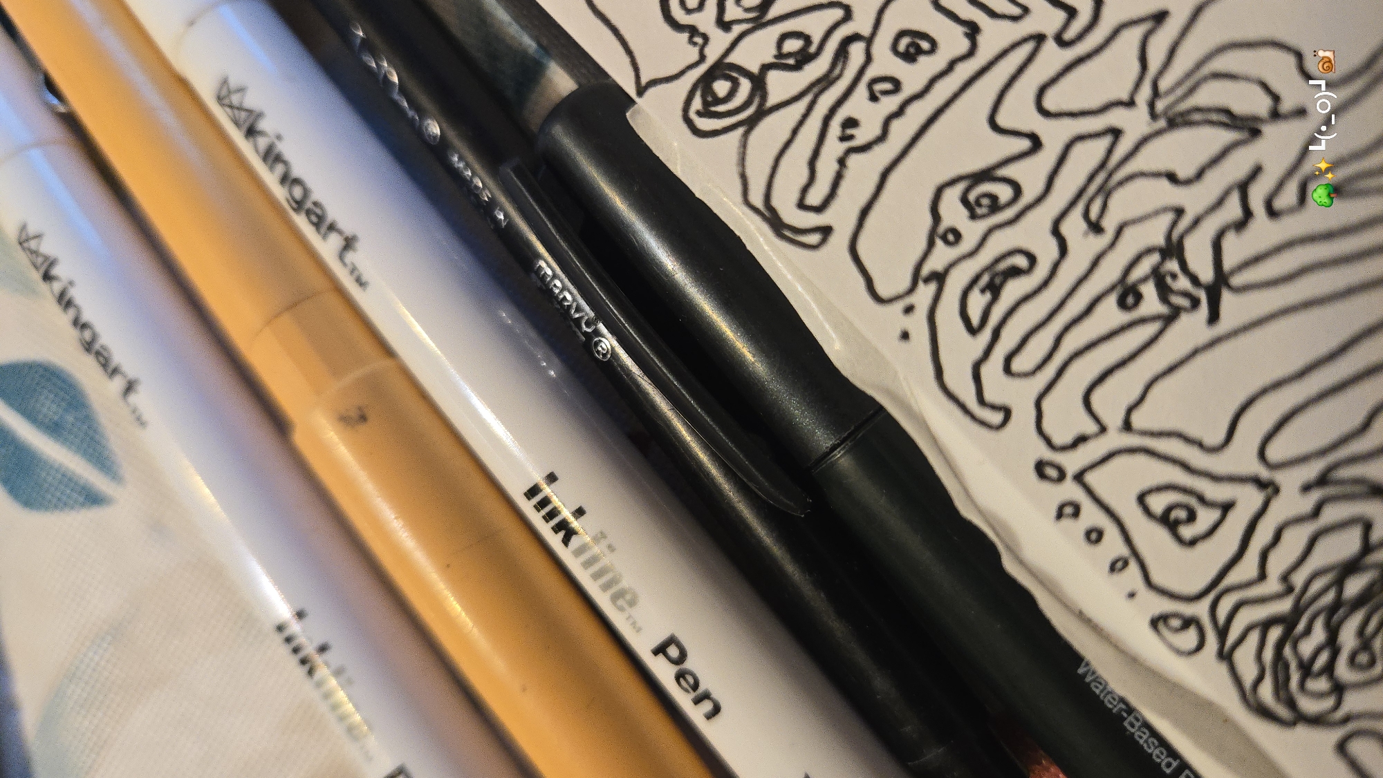 This close-up image captures various art supplies alongside a piece of paper displaying an abstract drawing. Several pens or markers are arranged diagonally across the frame. On the left, two white pens are visible, both branded with "kingart™". An orange marker lies between them and a black pen. The black pen, which appears to be a fineliner, features "marvU®" on its clip and "InkLine™ Pen" printed on its body. Below the black pen, partially visible text reads "Water-Based" on what seems to be its packaging or an adjacent white surface. These art tools rest on a patterned fabric, possibly a tablecloth, which displays a faint blue leaf design on the far left.
A white piece of paper occupies the right side of the frame, showcasing an intricate black line drawing composed of organic, interconnected shapes and loops, resembling a complex doodle or part of a larger artwork. In the top right corner of the image, a social media-style overlay adds a collection of emojis: a snail (🐌), a camera (📸), a face emoticon (•_•) , a sparkling star (✨), and a sprout (🪴). The scene suggests an ongoing or recently completed artistic activity, likely indoors on a table or desk, under soft, ambient lighting. No people are visible.