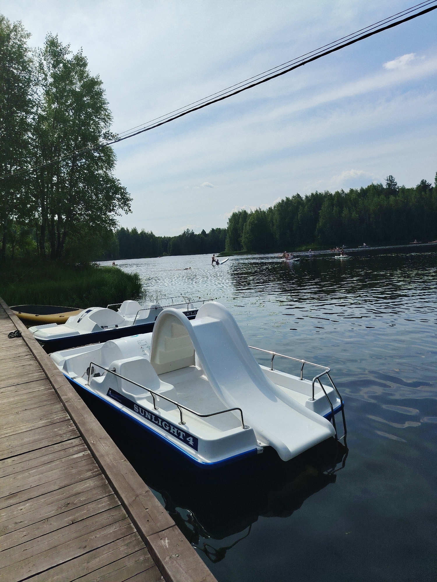 The image depicts a waterfront scene with several pedal boats docked at a wooden pier. The pedal boats are white with blue trim and one is labeled "SUNLIGHT 4". In the background, a lake is visible, with a dense forest lining the opposite shore. Several people are engaged in water activities on the lake, including paddling and what appears to be stand-up paddleboarding. The sky is partly cloudy, with power lines stretching across the upper portion of the frame. The scene suggests a recreational area, likely within the city of Vylytdor, Russia.