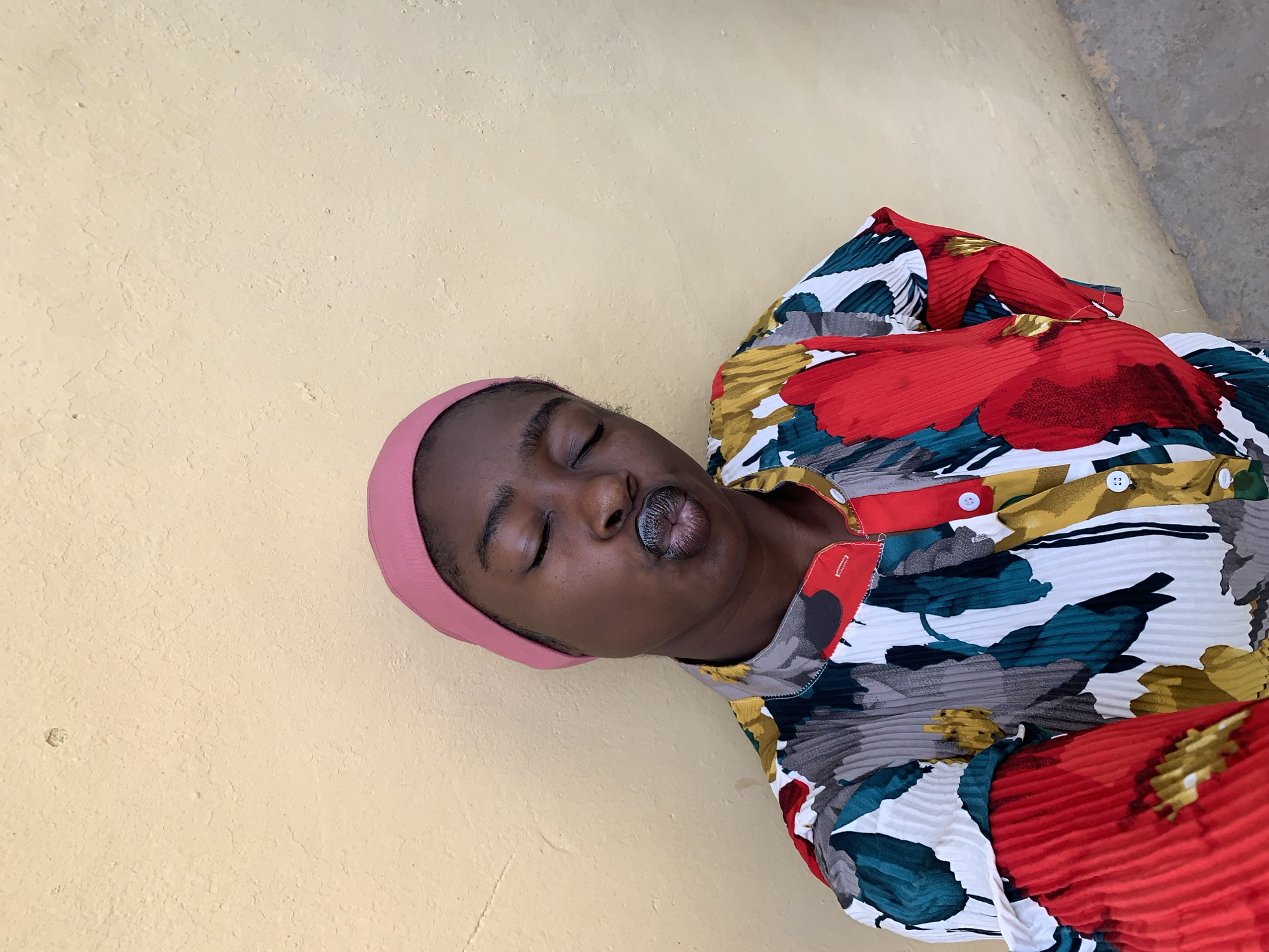 An individual with dark skin, closed eyes, and a pink head wrap lies on a painted, textured wall. The person is wearing a pleated blouse with a floral pattern in red, yellow, white, and teal. The location is Rafin Par, Nigeria.