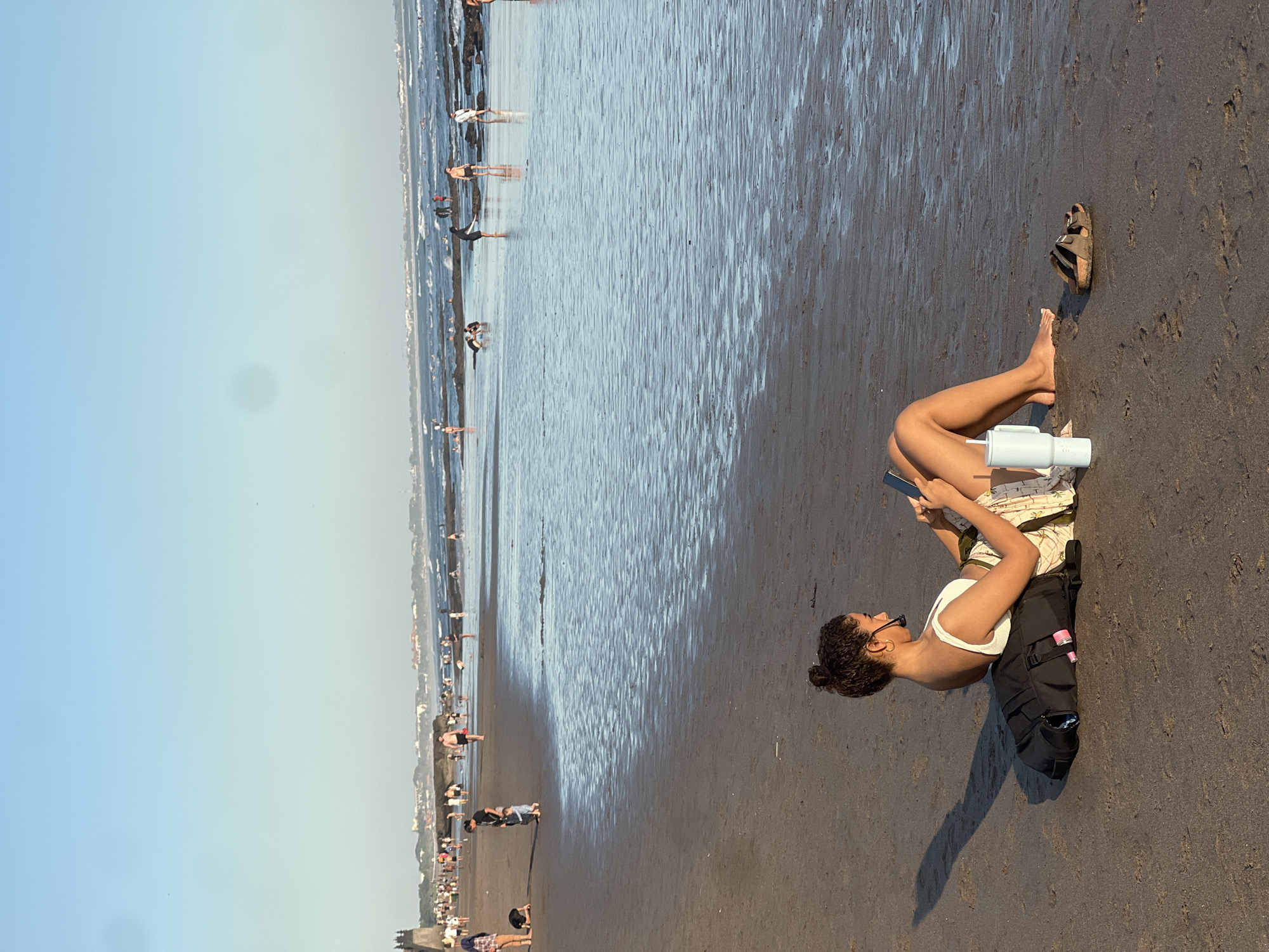 A medium shot captures a young woman sitting on a sandy beach in North Kuta, Indonesia, facing the ocean. She is positioned in the foreground, leaning back on a black backpack, and is wearing a white top and a patterned skirt. Her legs are extended towards the water, and she is holding a mobile phone. Next to her is a light blue insulated tumbler and a pair of brown sandals. In the background, numerous people are scattered across the beach and in the shallow water, some wading and others standing. The ocean has gentle waves, and the sky is clear and blue. The overall scene suggests a relaxed atmosphere on a sunny day.