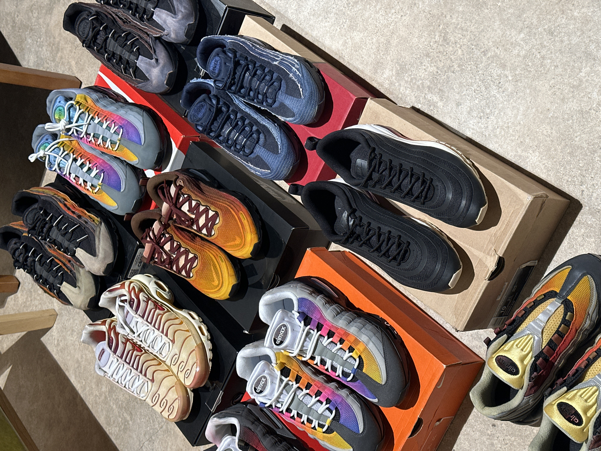 The image shows several pairs of Nike sneakers arranged on boxes. The sneakers are different models, including Air Max 95s and Air Max 97s, in various colorways. The boxes are stacked and scattered across what appears to be a concrete surface. The environment suggests an outdoor setting, potentially a sidewalk or loading dock.