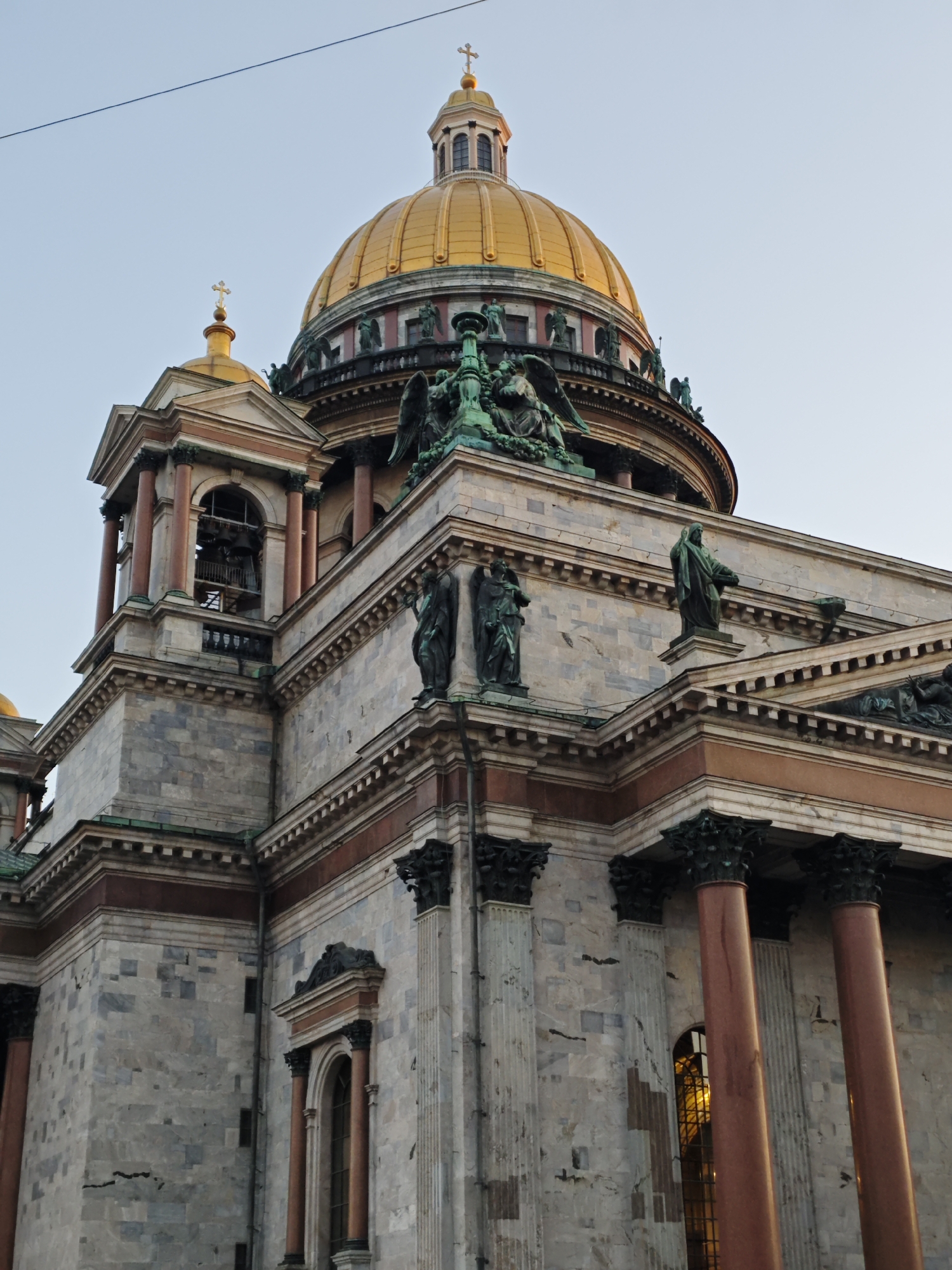 The image displays a low-angle exterior view of Saint Isaac's Cathedral, located in Saint Petersburg, Russia, against a clear, pale blue sky. The upper and mid-sections of the massive stone structure are visible.
A prominent, ribbed golden dome, topped with a smaller lantern structure and a cross, dominates the upper center. To its left, a smaller golden dome with a cross is partially visible. Below and to the left of the main dome, a square bell tower featuring four reddish-brown columns and arched openings reveals multiple dark-toned bells within. The building's facade is constructed from large, light-colored stone blocks, exhibiting a mottled grey and white pattern with occasional darker veins and lighter patches.
Multiple architectural layers are defined by reddish-brown cornices and entablatures. Dark, ornate capital designs are visible atop columns. Several large, dark-patinated statues, depicting winged figures and robed individuals, are positioned on various ledges and along the base of the main dome. One prominent winged figure is seated on a lower ledge on the left. Tall, arched windows, some appearing recessed and gridded, are visible on the lower right portion of the building, with one exhibiting an internal warm, golden reflection. A thin, dark linear object, likely a utility wire, crosses the sky in the upper left quadrant of the image. No people are present in the scene.