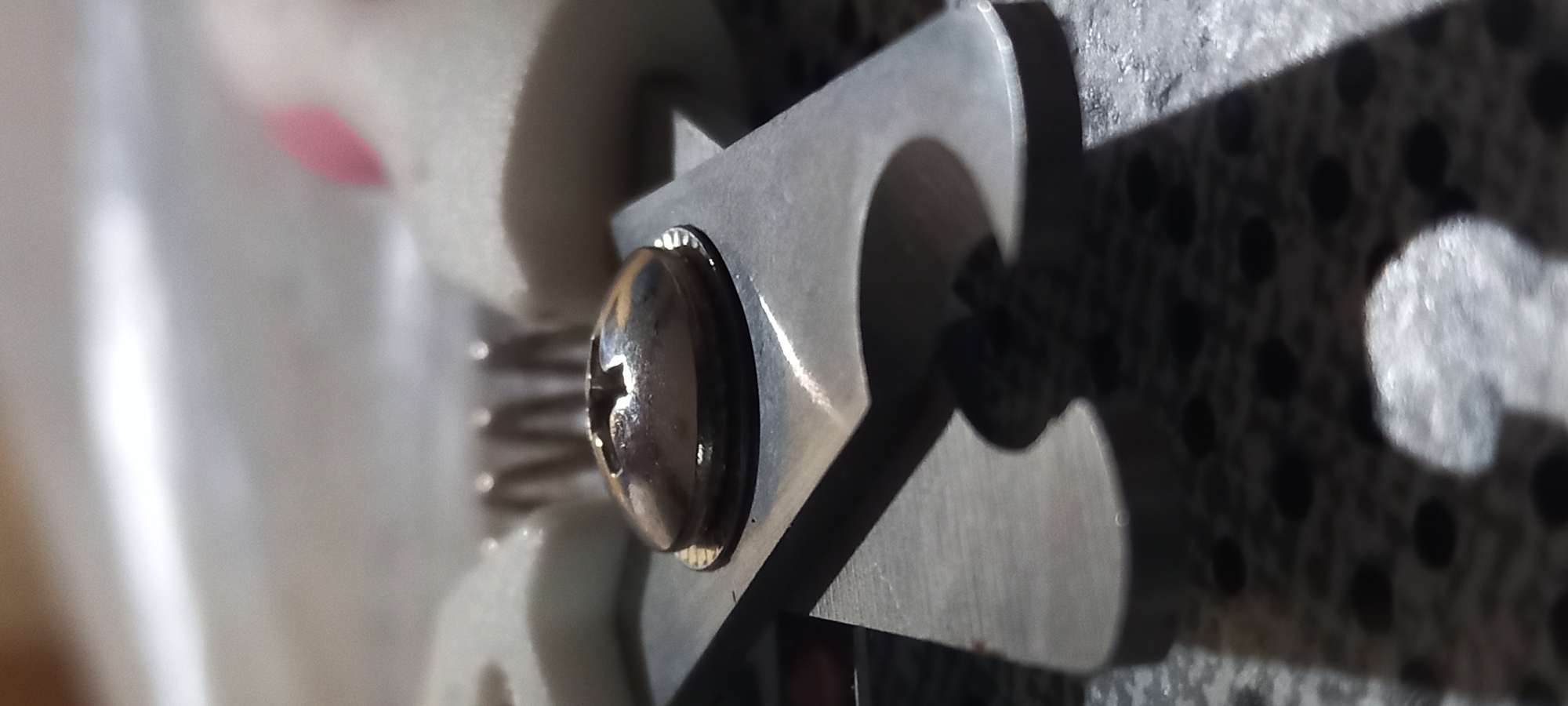 A close-up, macro-style photograph captures a metallic assembly of components under bright lighting. In the foreground, a prominent, shiny dark Phillips head screw is visible, securely fastened to a flat, silvery metallic piece. This metallic piece features a complex, angular, and curved profile, creating a shape that somewhat resembles a stylized numeral '2'.
Behind the screw and extending to the left, a coiled, silvery compression spring is present, positioned between the metallic piece and a blurry, light-colored, possibly white or off-white, object that appears to be made of plastic or ceramic.
To the right of the main metallic component, a sharp, dark shadow is cast onto a textured surface. This surface displays a consistent pattern of dark circular perforations or dots against a lighter background. The shadow itself mirrors the intricate outline of the metallic piece.
In the blurred upper-left background, a small, indistinct reddish-pink shape can be seen. The strong illumination creates distinct highlights and reflections on the metallic surfaces and sharp, well-defined shadows, suggesting a direct and intense light source, likely natural daylight. No human subjects, visible text, or explicit activity are depicted. The scene presents a static, detailed view of mechanical parts, offering no specific location cues within the frame for Kryvyi Rih, Ukraine.