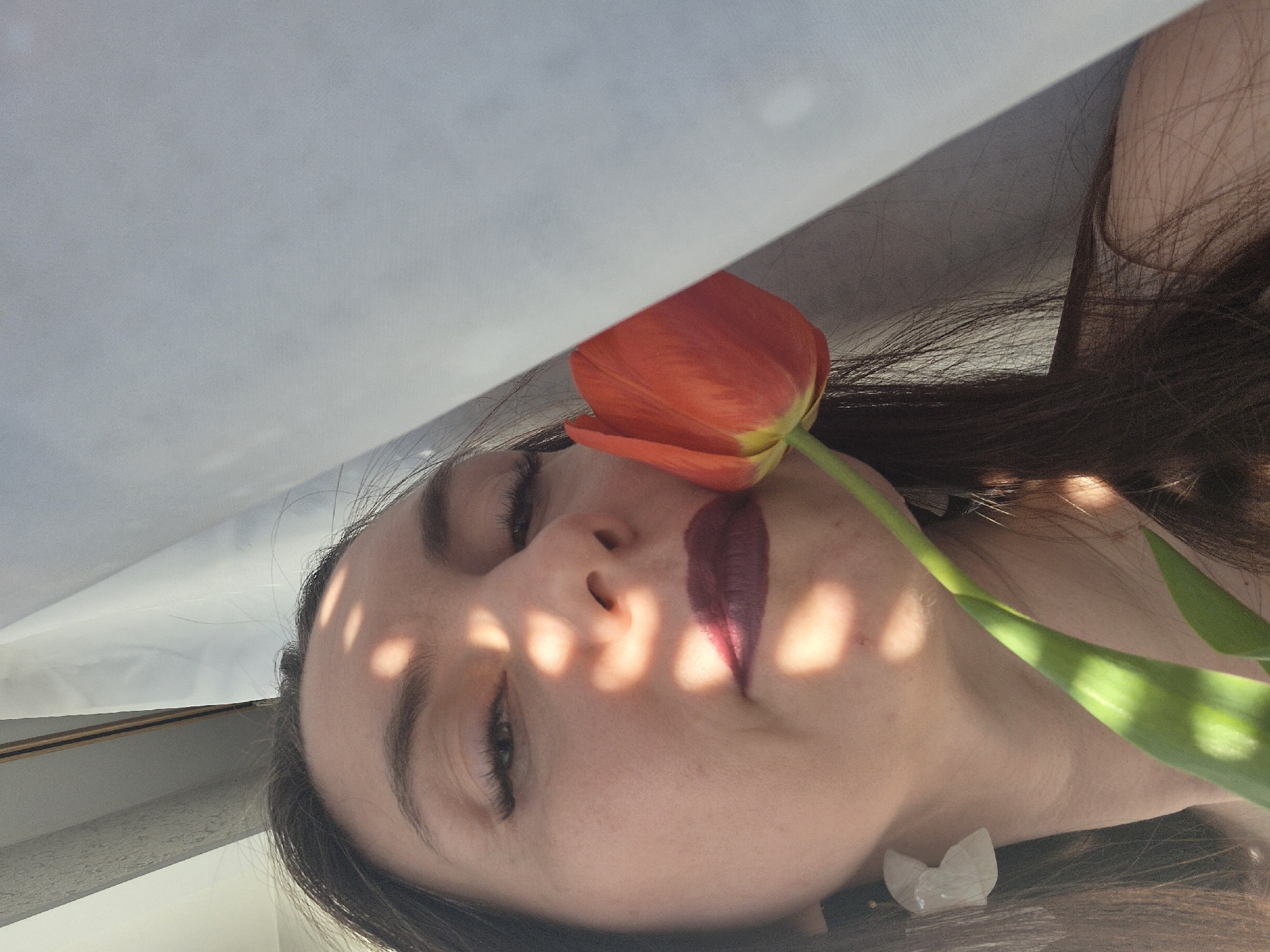 A close-up, rotated image shows a woman lying on her back, holding a red tulip near her face. She has dark hair and is wearing dark lipstick. Sunlight filters through an object, casting round shadows across her face. She appears to be wearing an earring that resembles a crystal. The background is blurred and unclear. Location and city are unknown.