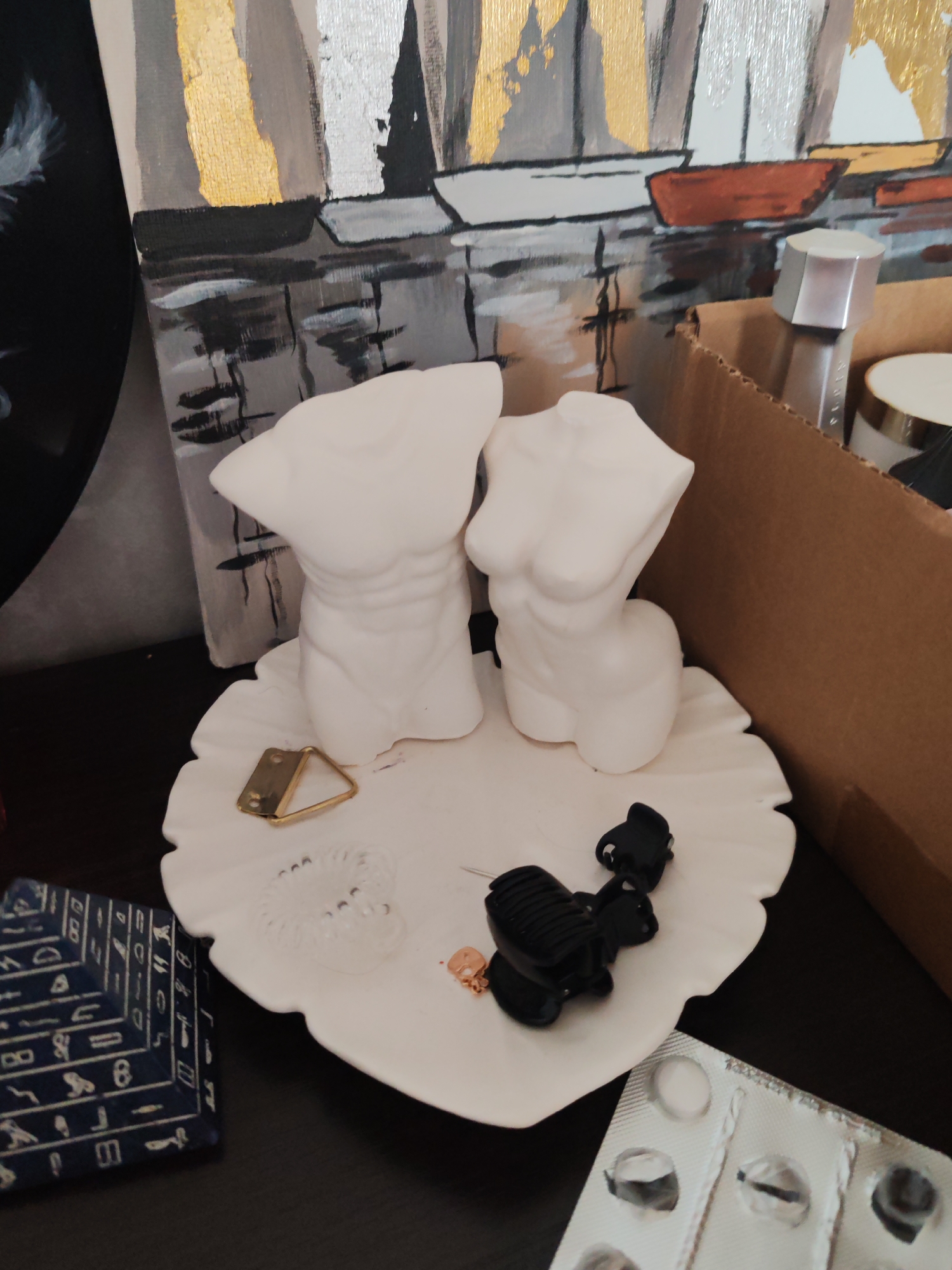 The image shows a still life composition. Two white, torso-shaped sculptures, one male and one female, are positioned on a white, ruffled plate. The plate also holds a gold-colored triangle hanger, a clear elastic hair tie, a small gold object, and two black hair clips.
To the left of the plate is a dark blue, pyramid-shaped object with hieroglyphic-like symbols on its sides. To the right, there's a partially empty blister pack of pills.
In the background, a painting depicting boats on water is visible. Behind the sculptures and plate is a cardboard box containing beauty products, including a silver-topped bottle and a jar.
The image doesn't provide any information that would indicate a location in Petropavlovsk-Kamchatsky, Russia, or any other specific geographic location.