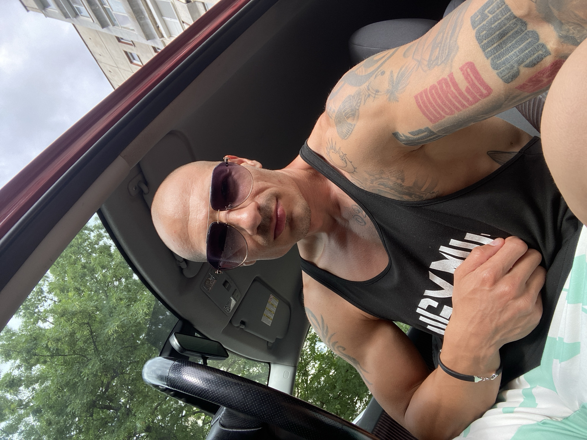 A bald, tattooed man wearing sunglasses and a black tank top sits in the driver's seat of a car. He is wearing a black bracelet on his left wrist. The car's sunroof is open, revealing trees and a cloudy sky. In the background, a multi-story building is visible. The man appears to be in the city of Pleven, Bulgaria.
