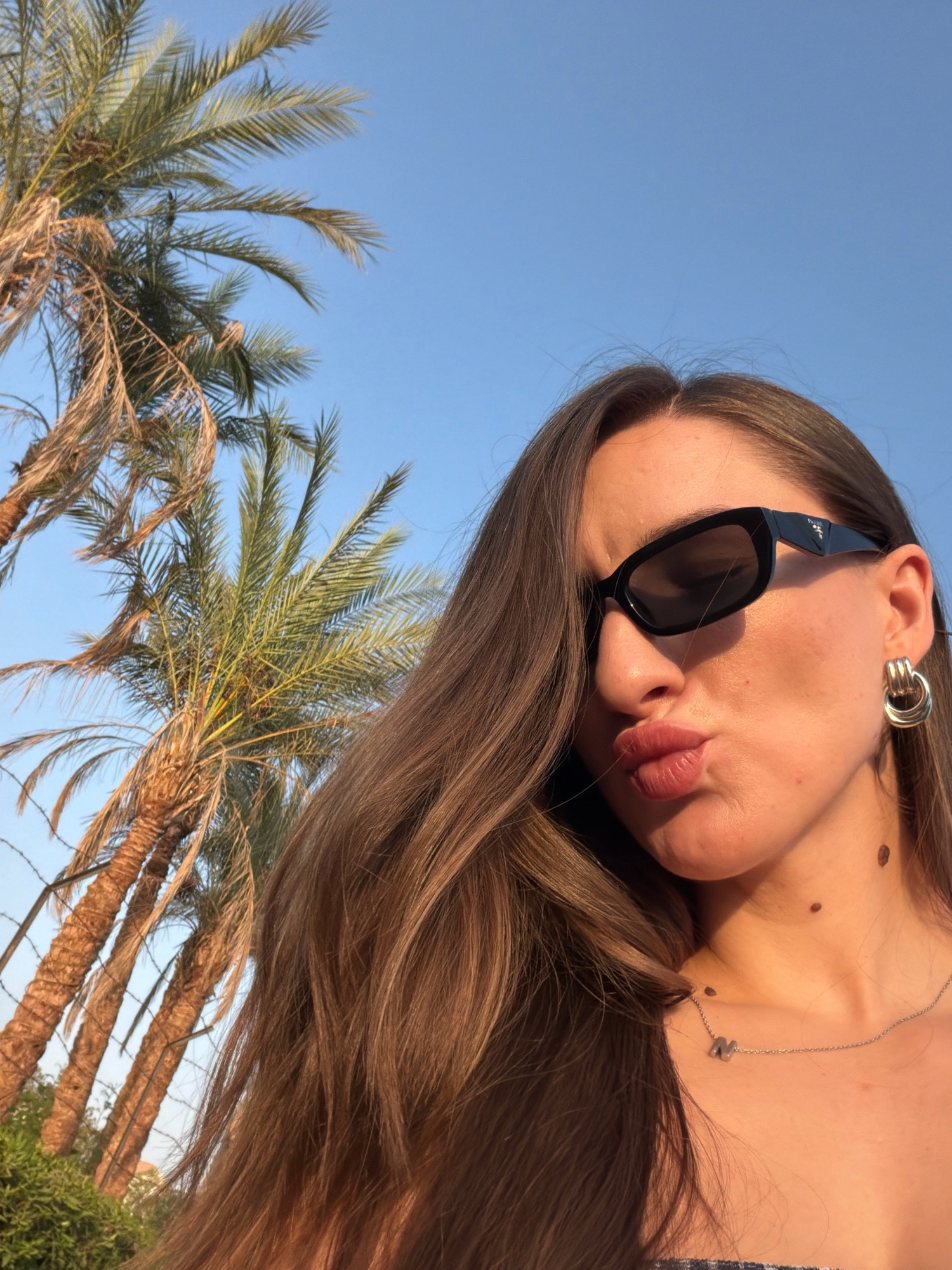 In Sharm El Sheikh, Egypt, a young woman with long, brown hair and sunglasses poses for a selfie against a clear blue sky, with palm trees and a wire fence visible in the background. She is wearing a gold hoop earring and a silver necklace with an "M" pendant. Her lips are pursed, and she is looking towards the camera. The lighting suggests it is daytime, likely late afternoon given the warm glow on her skin.