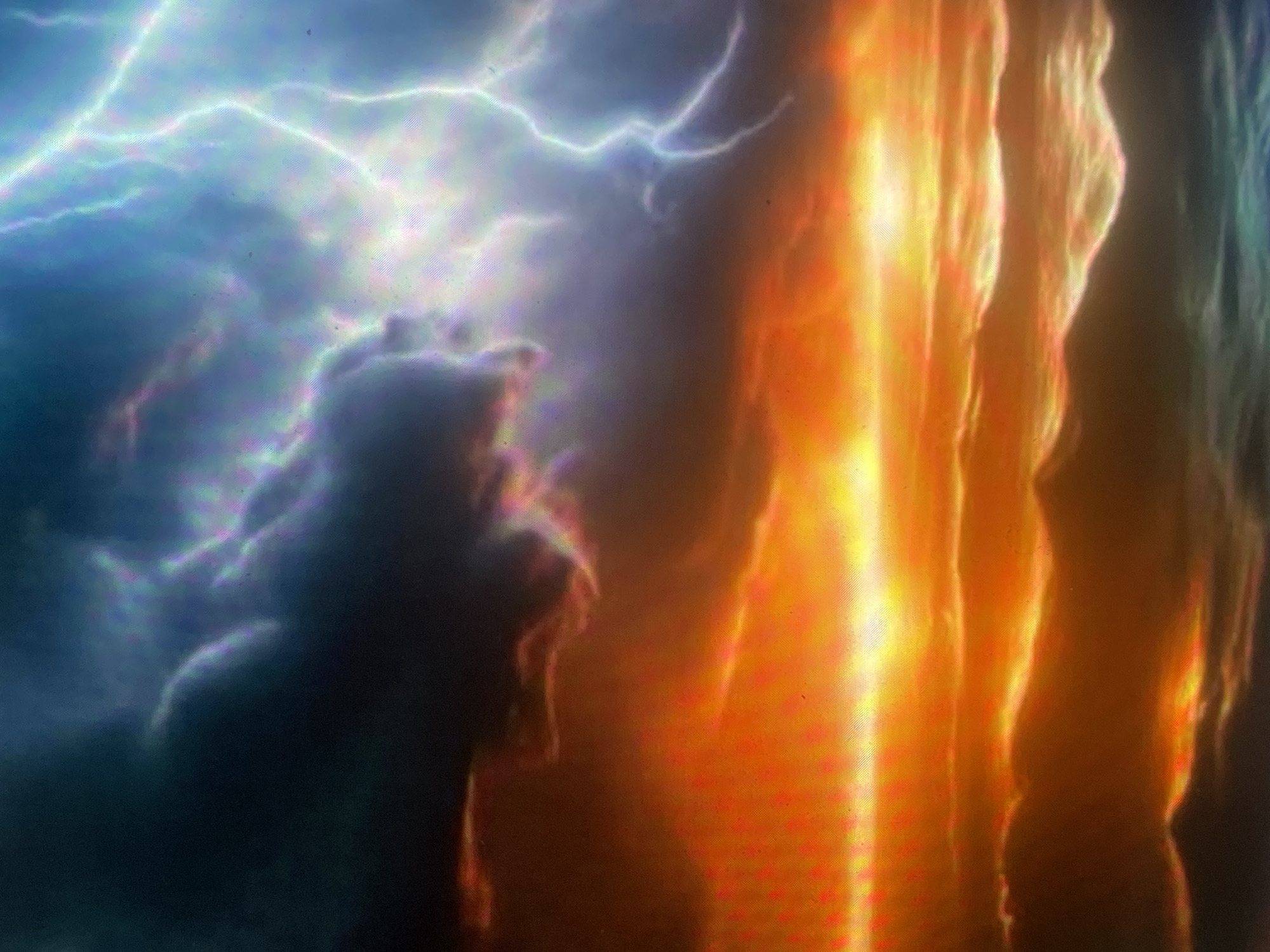 The image depicts a dramatic and highly stylized atmospheric scene, presented as being located in Zelenyi Hai, Ukraine. However, no specific geographical landmarks or structures are present to confirm this location visually.
The scene is divided into two contrasting halves, both horizontally and vertically. The upper portion represents the sky, and the lower portion represents a body of water, likely an ocean or sea.
In the upper-left quadrant, dark, heavy clouds dominate, showing subtle internal light effects with pixelated purple, green, and blue hues. The upper-right quadrant features intense, branching white and light-blue lightning strikes against a lighter, turbulent blue-gray sky. This area also exhibits noticeable pixelation and chromatic aberration, contributing to a vibrant, electric effect. A transitional zone in the upper-center shows a blend of dark cloud mass and scattered, faint light.
The horizon line separates the sky from the water. On the left, the sky near the horizon glows with an intense, fiery orange and yellow, suggestive of a powerful sunset or other incandescent atmospheric event. This warmth gradually transitions towards the darker, cooler tones of the storm on the right.
The lower portion of the image shows turbulent water with distinct waves. On the left, the water reflects the vibrant orange and yellow light from the sky, with the crests of the waves appearing brightly illuminated. On the right, the water is a darker, deep blue-green, reflecting the stormy sky above, with subtle white highlights indicating wave patterns.
The overall composition creates a stark contrast between a warm, almost infernal scene on the left and a cool, electrically charged storm on the right. The entire image displays a fine, grid-like artifact and localized chromatic aberration (rainbow fringing) across various areas, particularly visible in the brighter and darker sections, suggesting the image may be a photograph of a digital screen or a heavily processed digital artwork. No human figures, animals, or identifiable man-made objects are present in the scene.