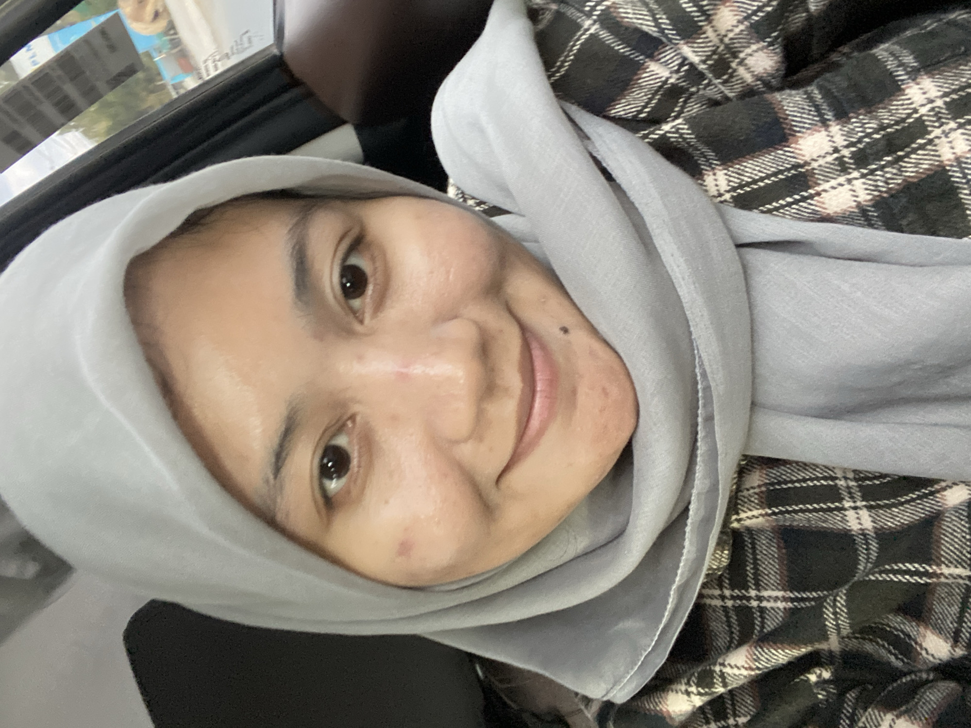 A close-up, selfie-style photograph shows a young woman wearing a light gray hijab and a dark plaid shirt. She is seated inside a vehicle, as indicated by the presence of a car door and window in the background, which offers a blurred view of an outdoor urban environment. The woman has light brown skin, with visible pores and a few blemishes on her face. Her eyes are dark brown and she is smiling gently. A small mole is visible on her chin. The lighting suggests it is daytime.