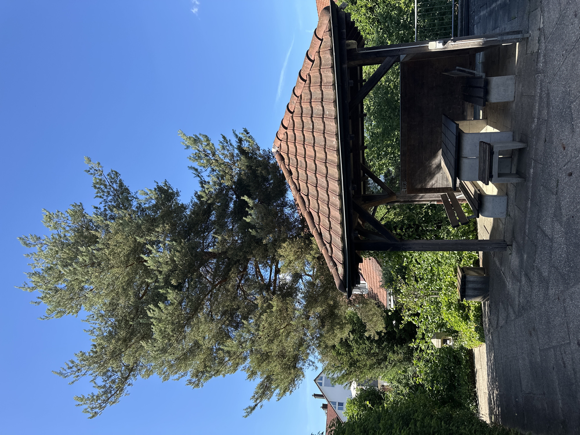 The original image shows a gazebo with a wooden structure and a tiled roof in Lufingen, Switzerland. Inside the gazebo, there is a stone table with wooden planks and two stone benches with wooden seats. To the left of the gazebo, there is a concrete bin. A large pine tree stands behind the gazebo, with its branches extending towards the clear blue sky. In the background, residential buildings with red-tiled roofs and lush green trees are visible. The ground is paved with concrete slabs, and shadows from the gazebo and trees are cast on the ground.