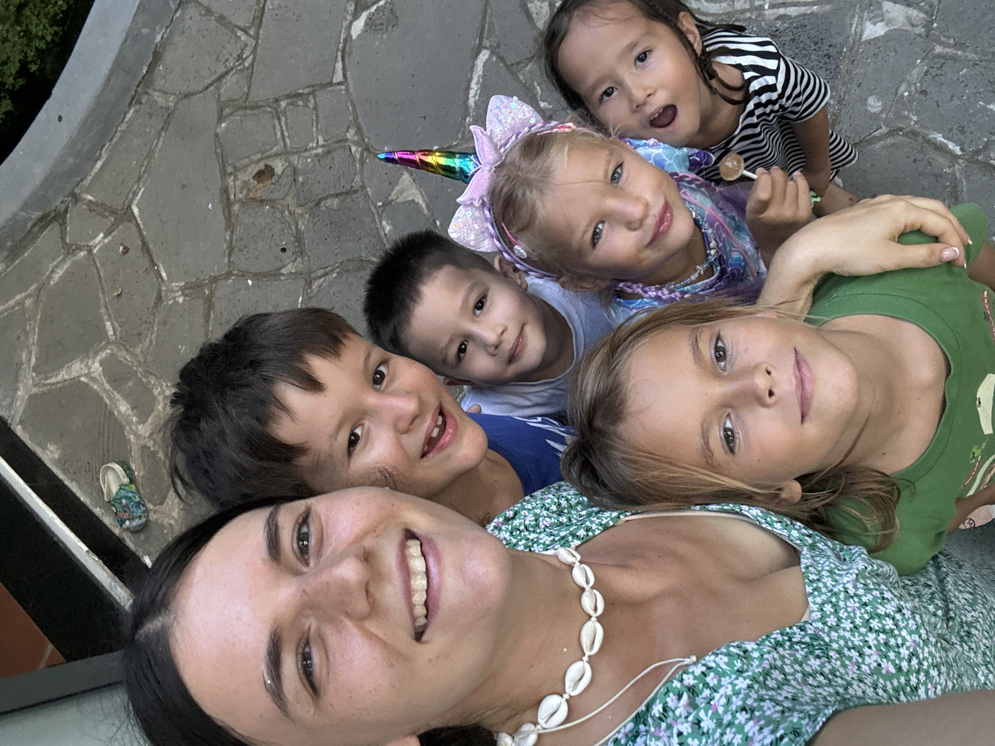A selfie photograph depicts a group of six individuals, likely a woman and five children. The woman, with dark hair and a green floral dress adorned with a shell necklace, smiles at the camera. Surrounding her are five children, all looking up at the camera. One child wears a unicorn headband, while another holds a lollipop. The setting appears to be outdoors on a paved surface, possibly a patio. A sandal is visible in the corner of the image. The location is not identifiable as Mũi Né, Vietnam, from the image alone.