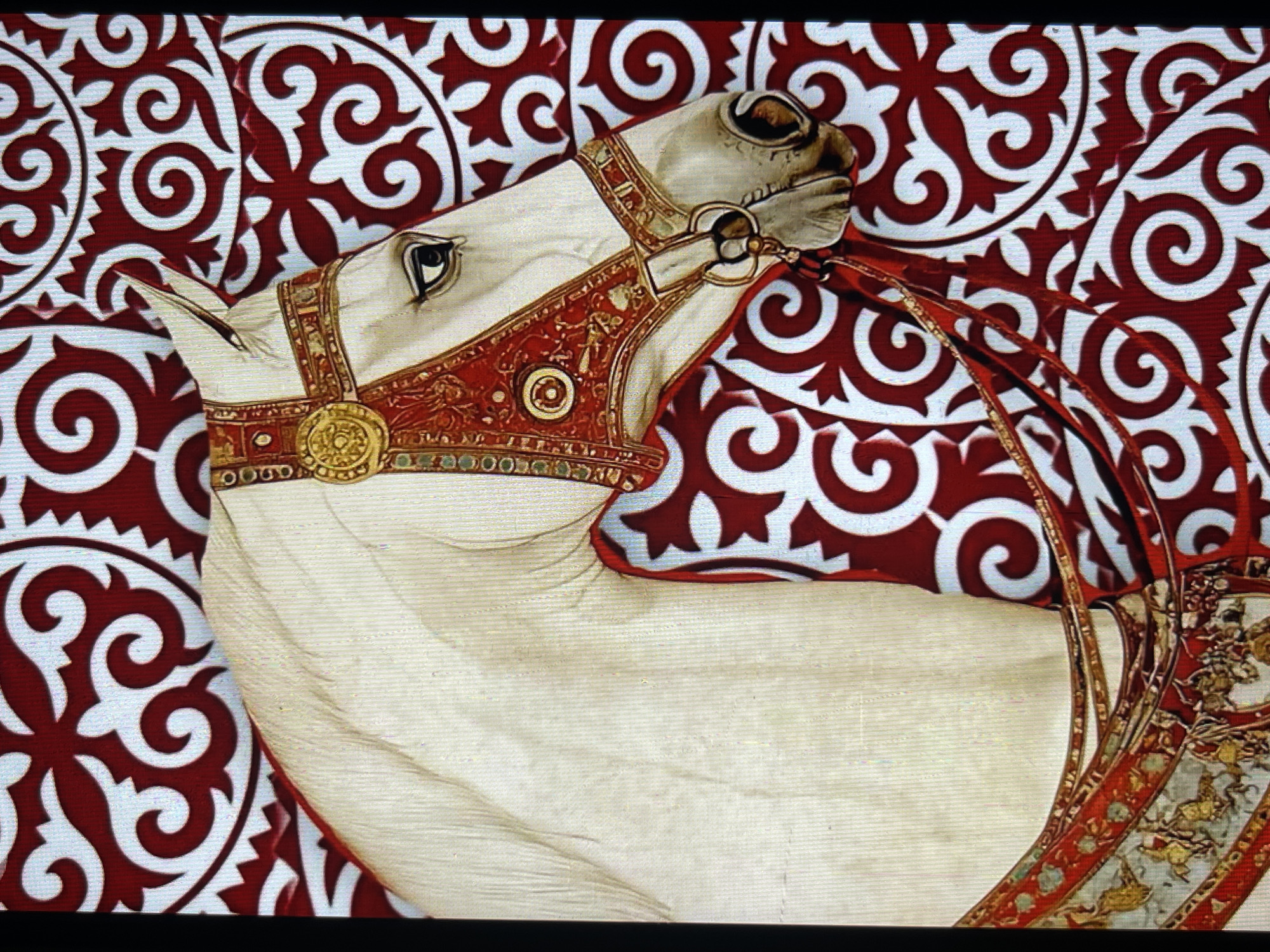 This is a close-up, head-and-shoulders shot of a white horse adorned with ornate red and gold bridlework and decorative trappings. The horse's head is turned to the right, and it appears to be looking forward. The background consists of a vibrant red and white patterned fabric, possibly a rug or tapestry, featuring swirling, symmetrical designs. The image is a detailed depiction of the horse and its elaborate attire against a decorative backdrop. The location is identified as Bishkek City, Kyrgyzstan.