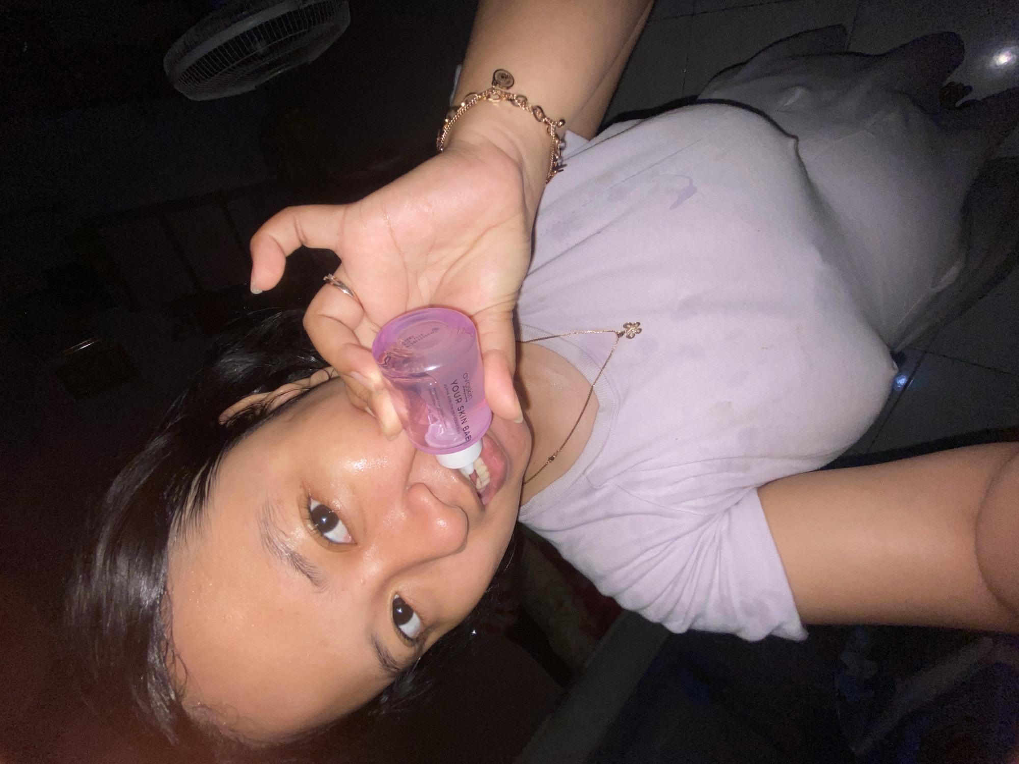 In a dimly lit room, a young woman with dark hair pulled back, showcasing sweat glistening on her forehead and neck, holds a small, pink, cylindrical bottle to her mouth. The bottle has text printed on it that reads "YOUR SKIN BABE" and "OVOGOD". She wears a light purple t-shirt, also damp with sweat, and a delicate gold necklace with a butterfly pendant. On her right wrist, she sports a rose gold bracelet with charms. Her gaze is directed at the camera, and her teeth are slightly visible as she grips the bottle. In the background, a standing fan is partially visible on the right side of the frame. The floor appears to be tiled. No specific location within Gelam, Indonesia is identifiable from the image.