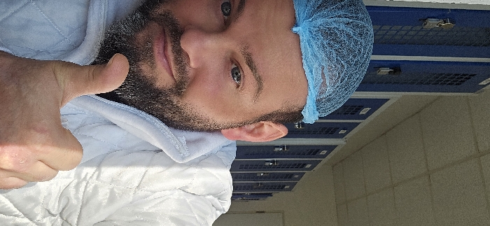 A male individual is depicted in a medium-close-up, taking a self-portrait. He has fair skin, blue eyes, and a full dark beard with visible grey strands. He wears a light blue disposable hairnet over his head and a white, quilted jacket or smock. His lips form a slight smile as he looks directly at the camera. His right hand, positioned in the lower center of the frame, is raised in a "thumbs up" gesture. The background consists of a row of dark blue lockers or storage units, each featuring a lighter blue mesh or gridded panel on its door. Several units exhibit silver-colored padlocks or similar securing mechanisms. The ceiling is light-colored and paneled. The environment appears to be an interior space, likely a changing room or preparation area within a facility requiring specific hygiene protocols. The specific location within Delbrück, Germany, cannot be determined from the visual content of this image.