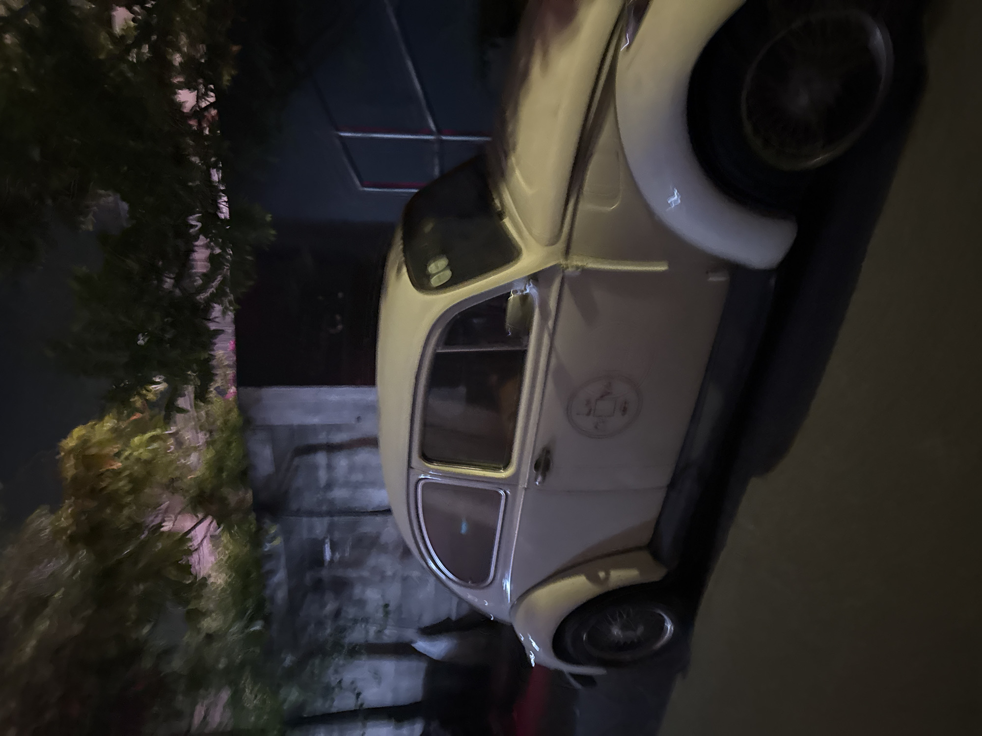The image shows a light-colored Volkswagen Beetle parked on a street at night, possibly in Hanoi, Vietnam. The car is positioned on the right side of the frame, facing towards the right. The environment is dark, with some trees and foliage visible in the background. There is a wall or building behind the car, and some light appears to be emanating from behind the trees. The car has a circular emblem on its side. The street surface is visible in the foreground, and it appears to be paved. There are no people clearly visible in the image. The overall impression is of a quiet, nighttime street scene in an urban setting.