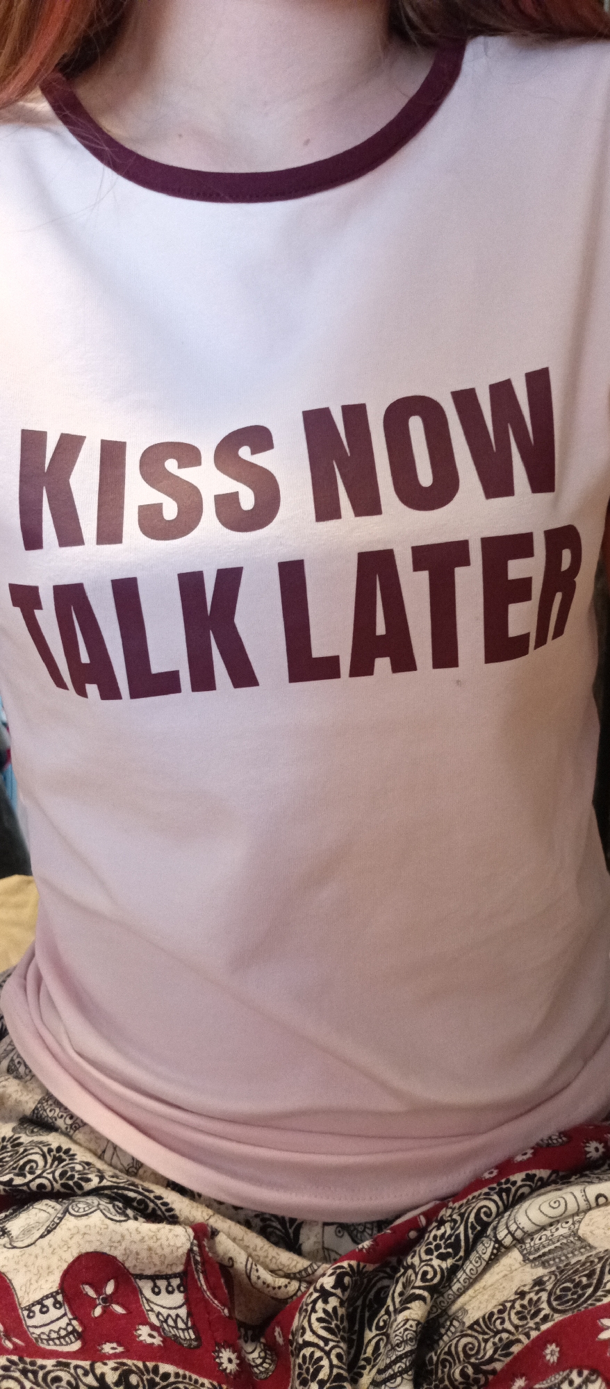 A close-up shot shows the upper torso of an individual wearing a light pink t-shirt with maroon trim around the collar. The shirt has the text "KISS NOW TALK LATER" printed on it in a dark maroon color. Below the shirt, a patterned fabric is visible, featuring a mix of cream, black, and red colors in an intricate design. The location is Unknown, Unknown.