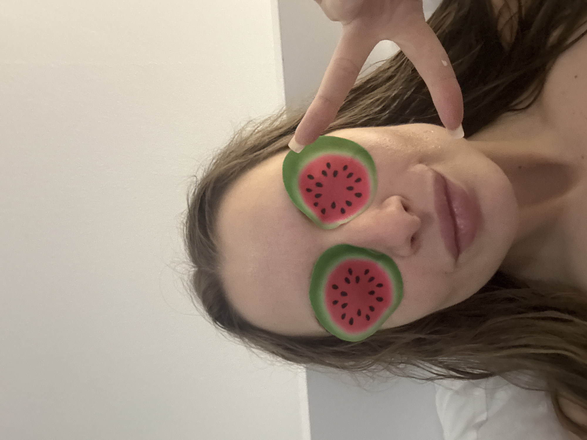 A woman is lying on her side, with two watermelon-shaped stickers covering her eyes. She is making a peace sign with her hand. The background is a plain white wall. There is no indication of the location being Budva, Montenegro.