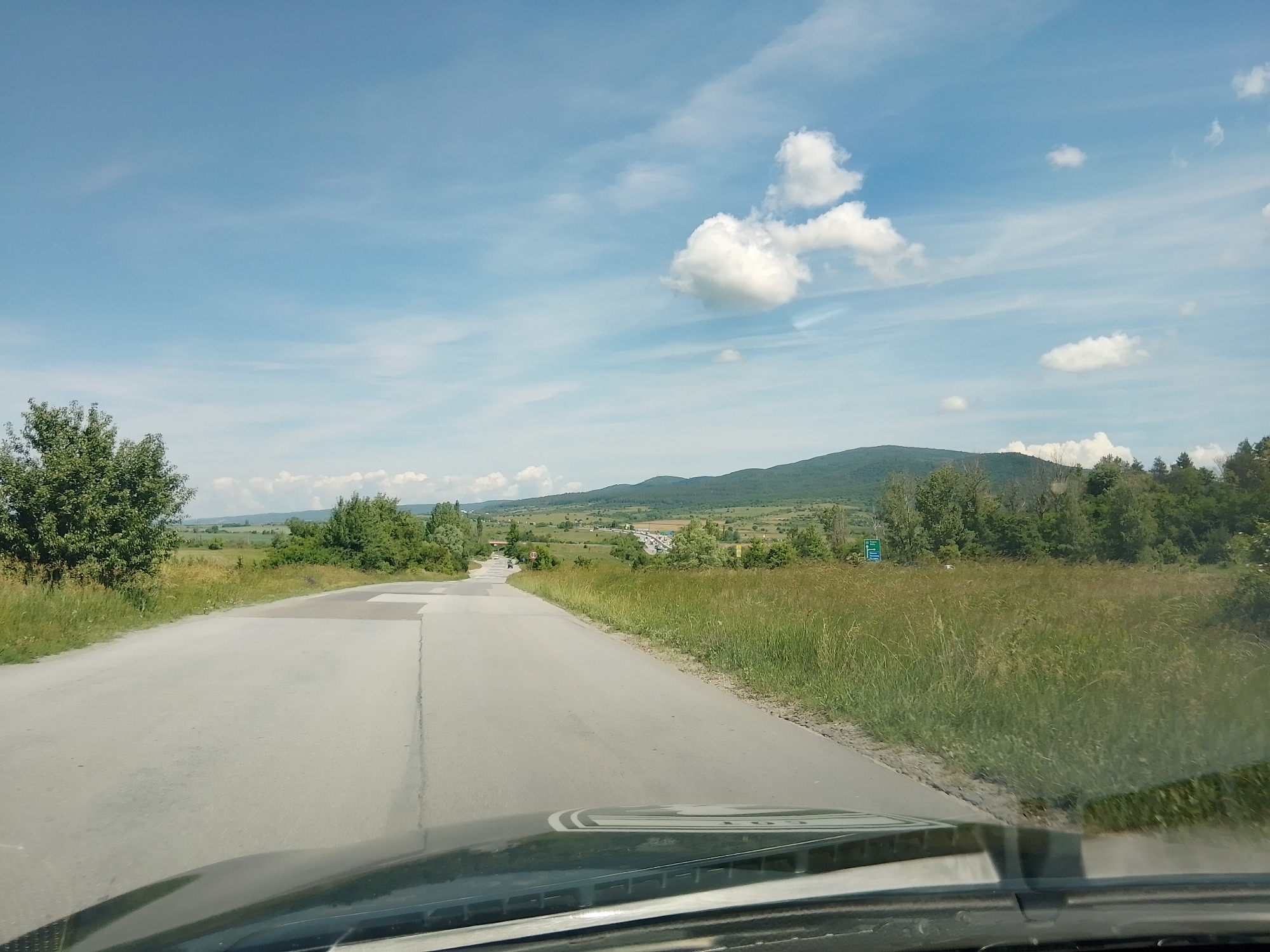 The image depicts a view from inside a vehicle, looking out at a paved road extending into a rural landscape under a bright, partly cloudy sky. The road appears to be two lanes, with a slight incline and a curve to the left in the distance. Lush green grass and trees line the sides of the road, with rolling hills and mountains visible in the background. A few scattered cumulus clouds are present in the expansive blue sky. The foreground shows the dashboard and windshield of the vehicle. There are no people or animals immediately visible in the scene. The overall impression is of a scenic drive through the countryside on a clear day.