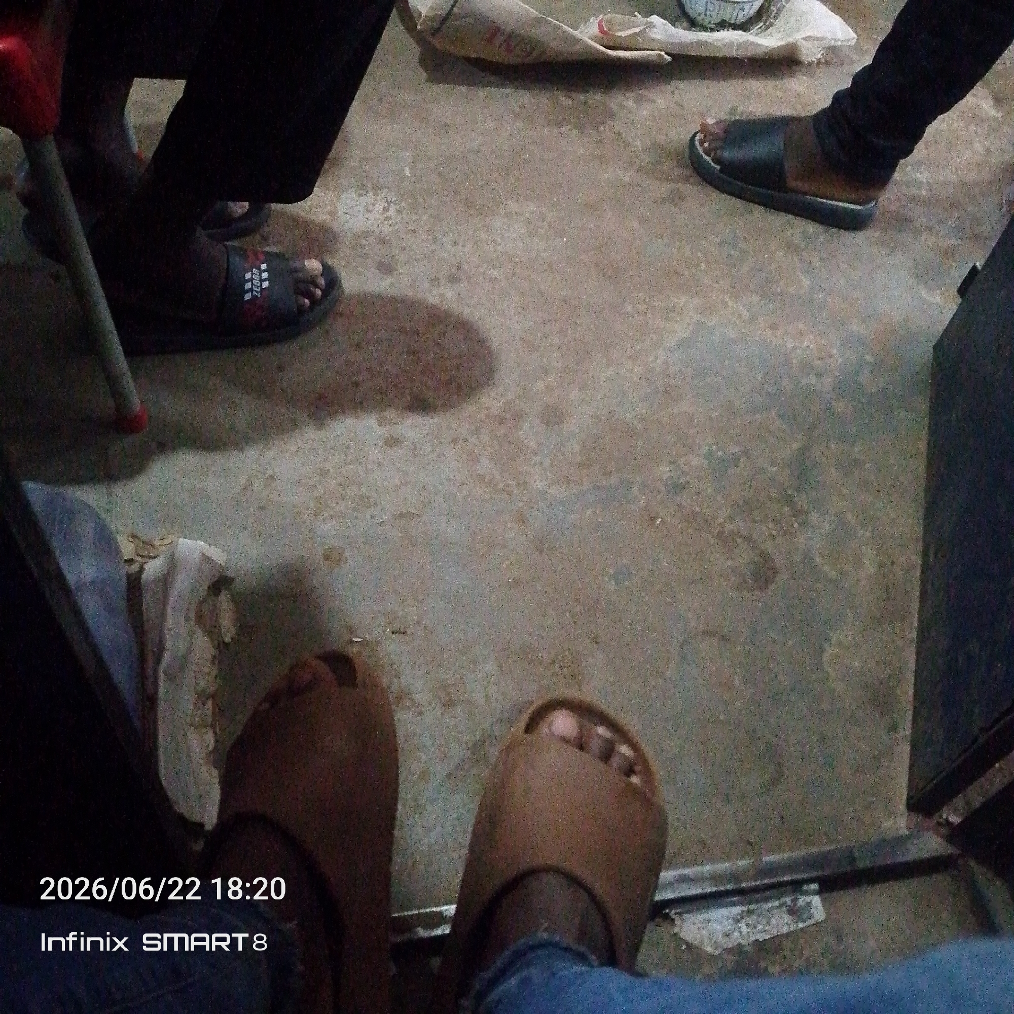 The image shows a low-angle view of several pairs of feet wearing sandals on a concrete floor. In the foreground, two feet are visible, each wearing a brown sandal. The left foot has a sandal that is mostly brown, while the right foot wears a light brown sandal with a darker strap. The background shows more feet, including one pair wearing black slides with red accents and another pair wearing black slides with a brown band. The floor is a rough concrete surface with some dark staining and lighter patches. There is a red object on the left side of the frame, which appears to be the leg of a chair or table. In the upper right corner, a dark, rectangular object is partially visible, possibly furniture. The lighting is dim, suggesting it might be evening or indoors. There is text overlay on the bottom left corner of the image. The text reads "2026/06/22 18:20" and "Infinix SMART 8".