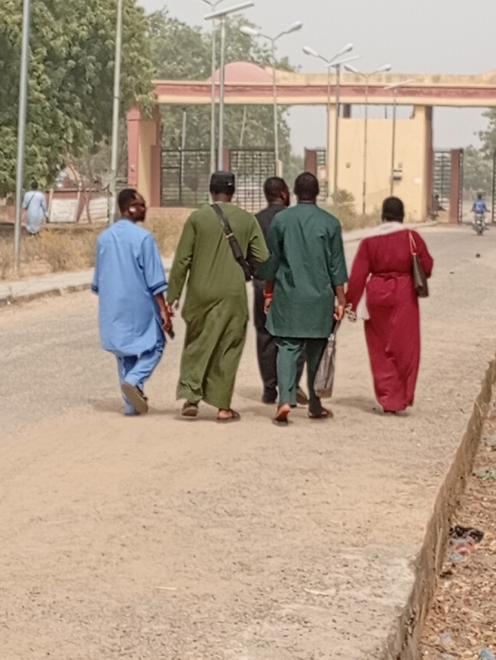 Five people walk away from the camera on a road leading towards a large gate. The individuals wear various colored robes: light blue, olive green, dark green, black, and maroon. The gate has a red domed roof and multiple arched openings. In the distance, a person rides a motorcycle. The sky is hazy. Trees surround the area and appear to be on either side of the road.
