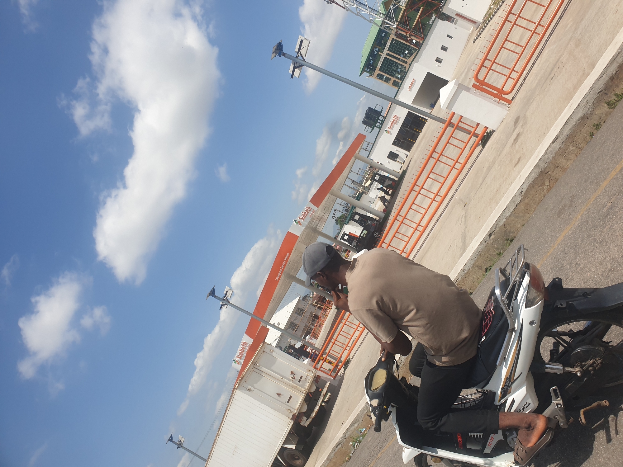 A medium, tilted, outdoor shot captures a man on a motorcycle in front of a Sunbeth Service Station, identified as Lokongoma Service Station in Lokoja, Nigeria. The man is wearing a gray cap, brown t-shirt, black pants, and brown sandals. He is seated on a white and black motorcycle. Behind him is the service station, and to his left is a white truck. The sky is blue with white clouds. Streetlights are visible in the background.