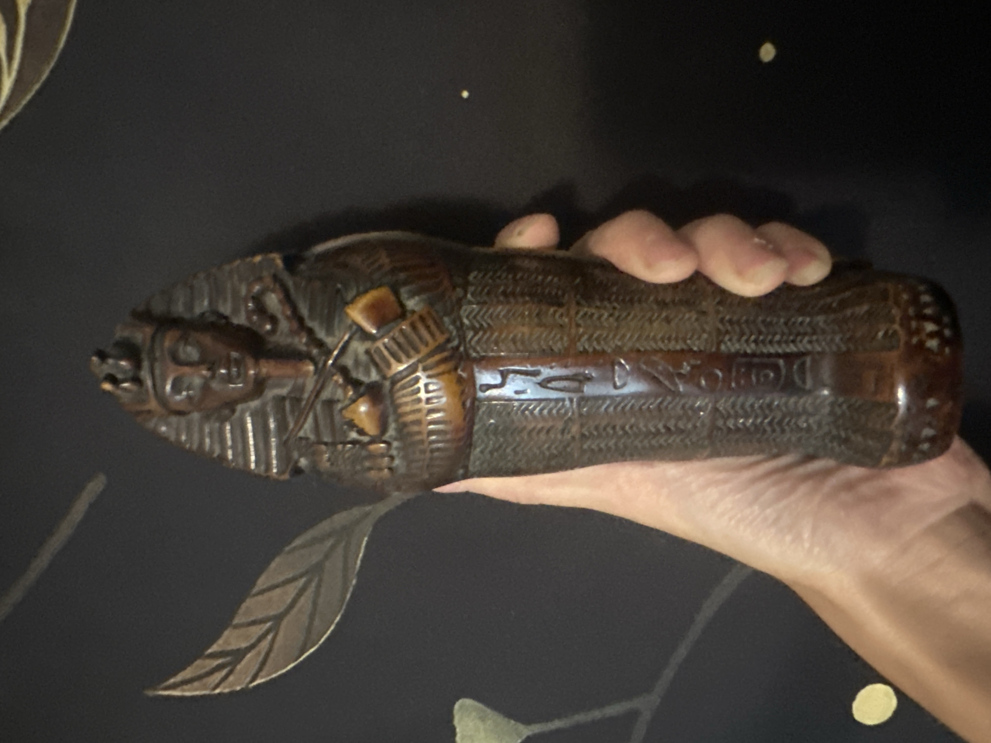 A hand holds a small, dark brown, carved object resembling an Egyptian sarcophagus with hieroglyphs. The object is detailed with a carved face wearing a nemes headdress, crossed arms with miniature tools, and textured body covering. The background is dark and appears to be a wall with a subtle pattern of leaves and dots. This image was taken in Togliatti, Russia.
