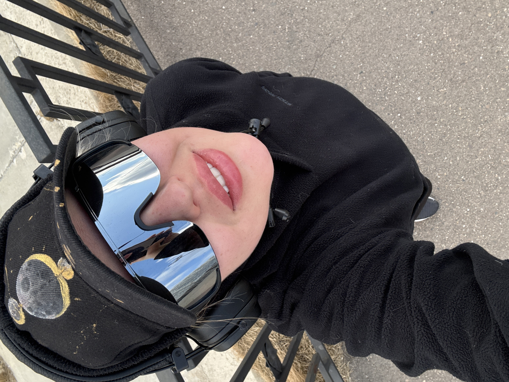 This close-up outdoor portrait, rotated counter-clockwise, features a person in Krasnoyarsk, Russia. Only the lower part of their face, from the nose down, is visible. They have lightly glossed lips, slightly parted to reveal white teeth.
The person is wearing large, futuristic-looking reflective wraparound sunglasses that mirror a clear, bright sky and a distant horizon. A black fleece jacket covers their torso, with a visible drawstring toggle and partially legible text that appears to read "STOLICHNYE" on the chest. Black over-ear headphones are worn, with strands of light-colored hair visible underneath. A black baseball cap is worn, decorated with a distinct grey and gold moon-like design on the side.
The background includes a black metal railing with horizontal bars positioned behind the person. Beneath the railing, dry, light brown grass is visible. The ground surface is paved, appearing to be asphalt or concrete with a speckled texture. The scene depicts a clear, bright daytime setting.