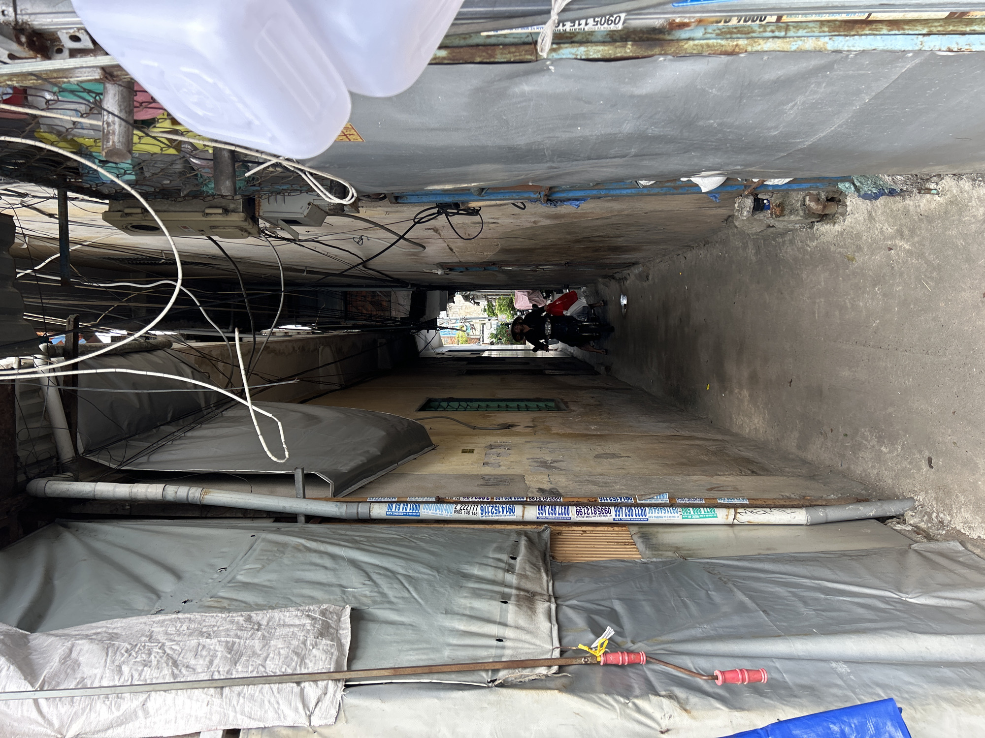 The image captures a narrow, enclosed alleyway or corridor in a dense urban setting, consistent with Nha Trang Ward, Vietnam. The perspective looks down the long, tight passage, which is flanked by tall, worn walls of concrete or stucco.
The alley's ceiling is a patchwork of various materials, including corrugated metal, tarps, and pipes, suggesting improvised roofing. Overhead and along the walls, a tangled network of black electrical wires and cables crisscrosses the space, with several small, grey electrical boxes attached. A large white plastic container hangs near the foreground on the left.
In the distance, at the far end of the alley, a person is visible on a scooter or motorcycle, seemingly navigating or waiting. Beyond them, a glimpse of brighter outdoor light, some greenery, and what appears to be laundry hanging on lines suggests an exit to a more open area. The lighting indicates it is daytime, with shadows dominating the alley but a brighter patch at the end.
Along the lower sections of both walls, large grey pipes run horizontally. The pipe on the right (from the perspective looking down the alley) is covered with numerous white stickers or labels featuring various Vietnamese phone numbers, such as "0914 152 116," "0977 297 699," "0935 812 199," "0933 052 466," and "0931 048 000," as well as the word "DURA" and other partially legible Vietnamese text. These likely serve as advertisements or contact information.
The floor is concrete, appearing aged and somewhat uneven. In the immediate foreground, grey tarpaulins cover the ground or lower wall sections, with a metal rod and a red-handled tool visible. The overall scene portrays a functional, lived-in urban environment characterized by improvisation and practical use of available space.
