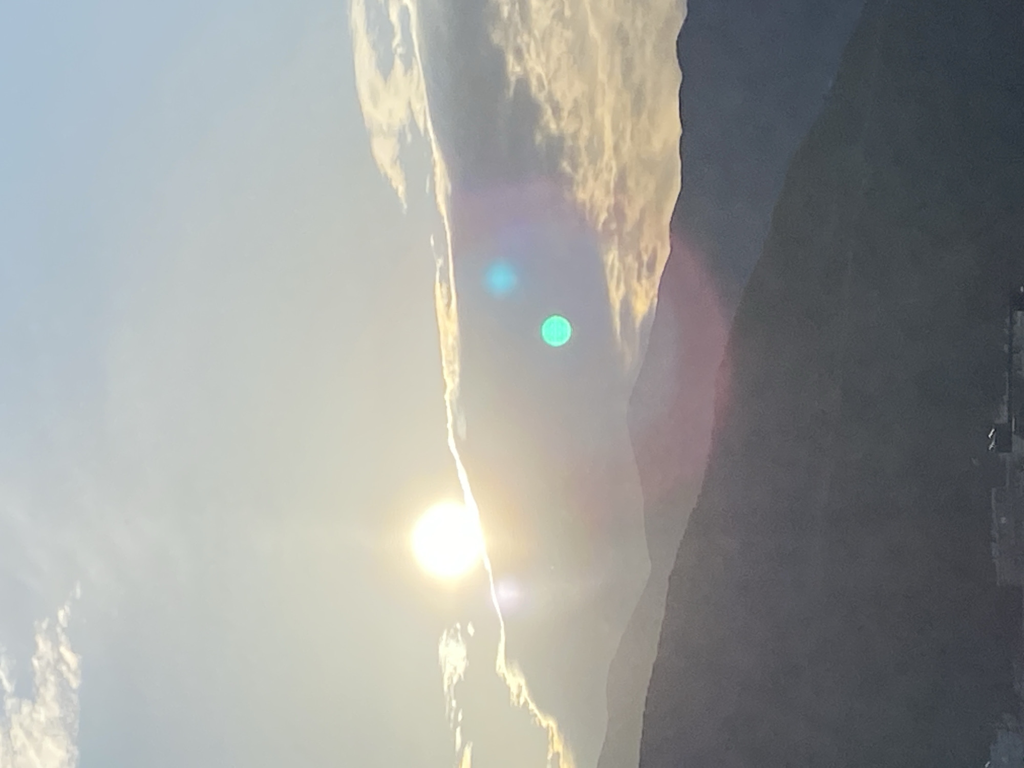 The image shows a landscape with mountains and the sun. The sun is a bright, circular shape located in the lower left portion of the image. There are clouds partially obscuring the sun. To the right of the sun are tall, dark mountains, taking up a large portion of the right side of the image. In the background, some buildings or structures are visible near the base of the mountains. The location might be Budva, Montenegro based on the landscape characteristics.