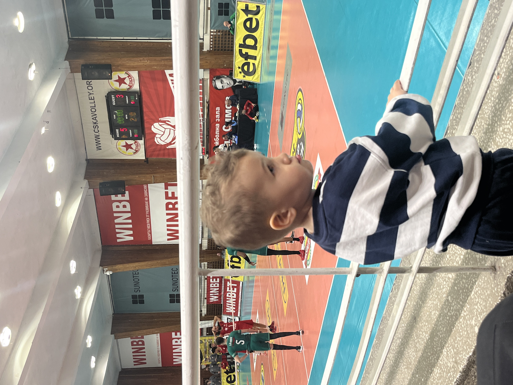 An indoor scene depicts a volleyball match from a spectator's perspective in the Vasil Simov Volleyball Hall, Sofia, Bulgaria.
In the immediate foreground, a young child, likely a toddler, is positioned with their back mostly to the viewer, observing the volleyball court. The child wears a dark blue and white horizontally striped long-sleeved shirt and dark pants, with their right hand resting on a white metal railing composed of multiple horizontal bars.
Beyond the railing, a two-tone volleyball court is visible, featuring orange and blue sections with yellow and white boundary lines. Several male volleyball players are present on the court. Two players in green jerseys and black shorts are identifiable; one, labeled "L. BOJIDOV" with number "5" on his back, stands with hands on his hips. Two players in red jerseys and shorts are also visible, one with hands on his head.
The background reveals the arena's interior. A light-colored ceiling contains multiple recessed circular light fixtures. The walls are paneled with vertical wooden strips interspersed with various advertising banners. Prominent red and white "WINBET" banners display Bulgarian text translated as "GAMBLING CARRIES A RISK OF DEVELOPING ADDICTION!" A large blue "SUNOTEC" banner is also visible. Above the court, a black electronic scoreboard displays "HOME 5," "TIME 00:00," "SETS 2:0," and "GUEST 3," flanked by two red star logos with "ЦСКА" (CSKA) and the web address "WWW.CSKAVOLLEY.ORG" above it. Other banners include "ВОЛЕЙБОЛНА ЗАЛА" (Volleyball Hall) and a large portrait of a man. An "efbet" banner is also displayed. Black speakers are mounted on the walls near the scoreboard, and a white security camera is mounted high on a wall. Several individuals, likely officials or staff, are seated at a table behind the court, with one person in a red tracksuit kneeling on the court near the table.