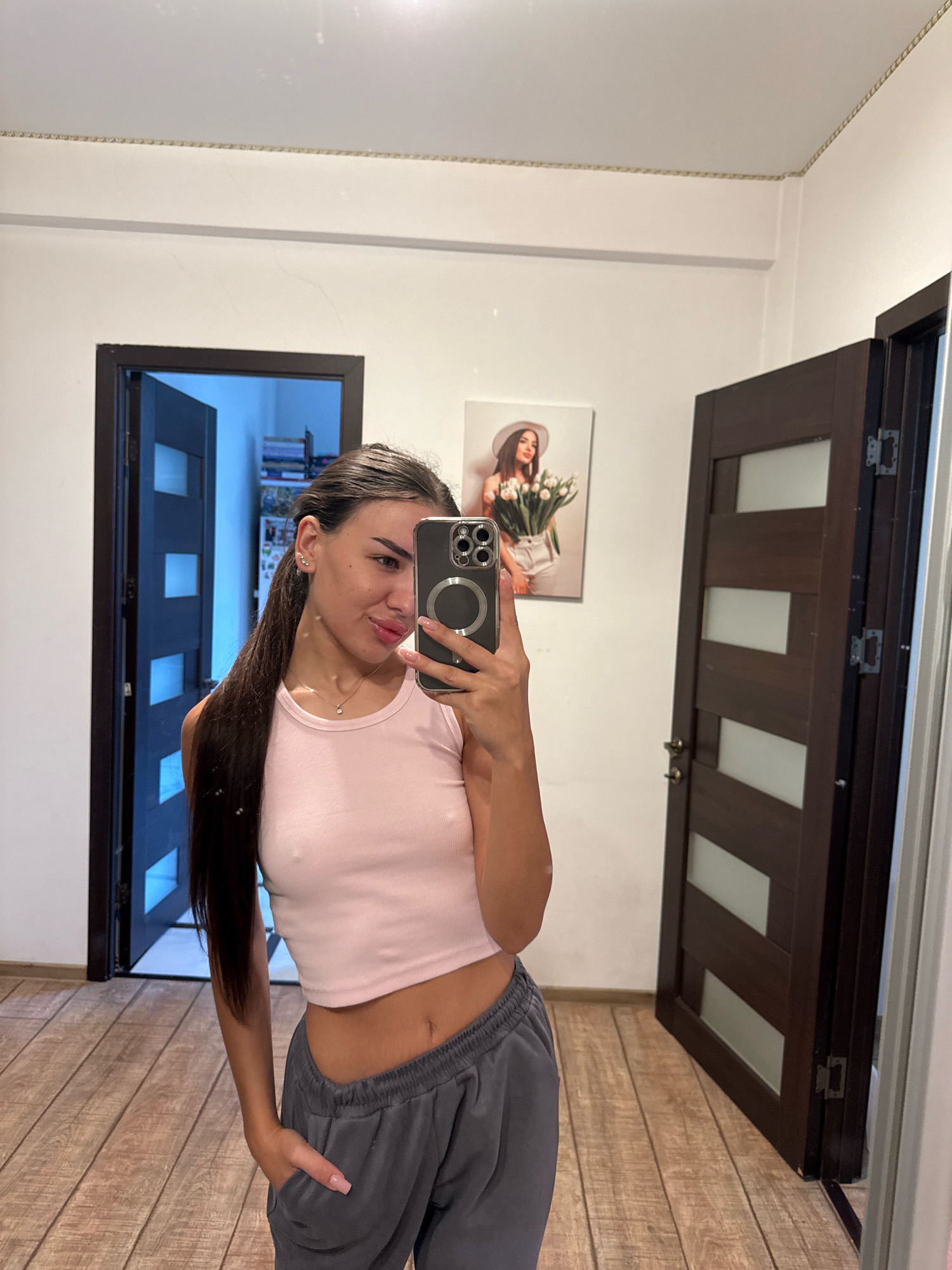 A young woman with long dark hair, wearing a pink tank top and grey sweatpants, takes a mirror selfie in what appears to be an apartment hallway. She is holding a smartphone in her right hand, and her left hand is in her sweatpants pocket. Behind her, there are two closed wooden doors, one with frosted glass panels. To the left of the woman, a door is ajar, revealing shelves with books and other items. On the wall to the right, a framed picture of a woman holding flowers is visible. The flooring is wooden. The overall impression is that of a casual moment captured within a residential setting.