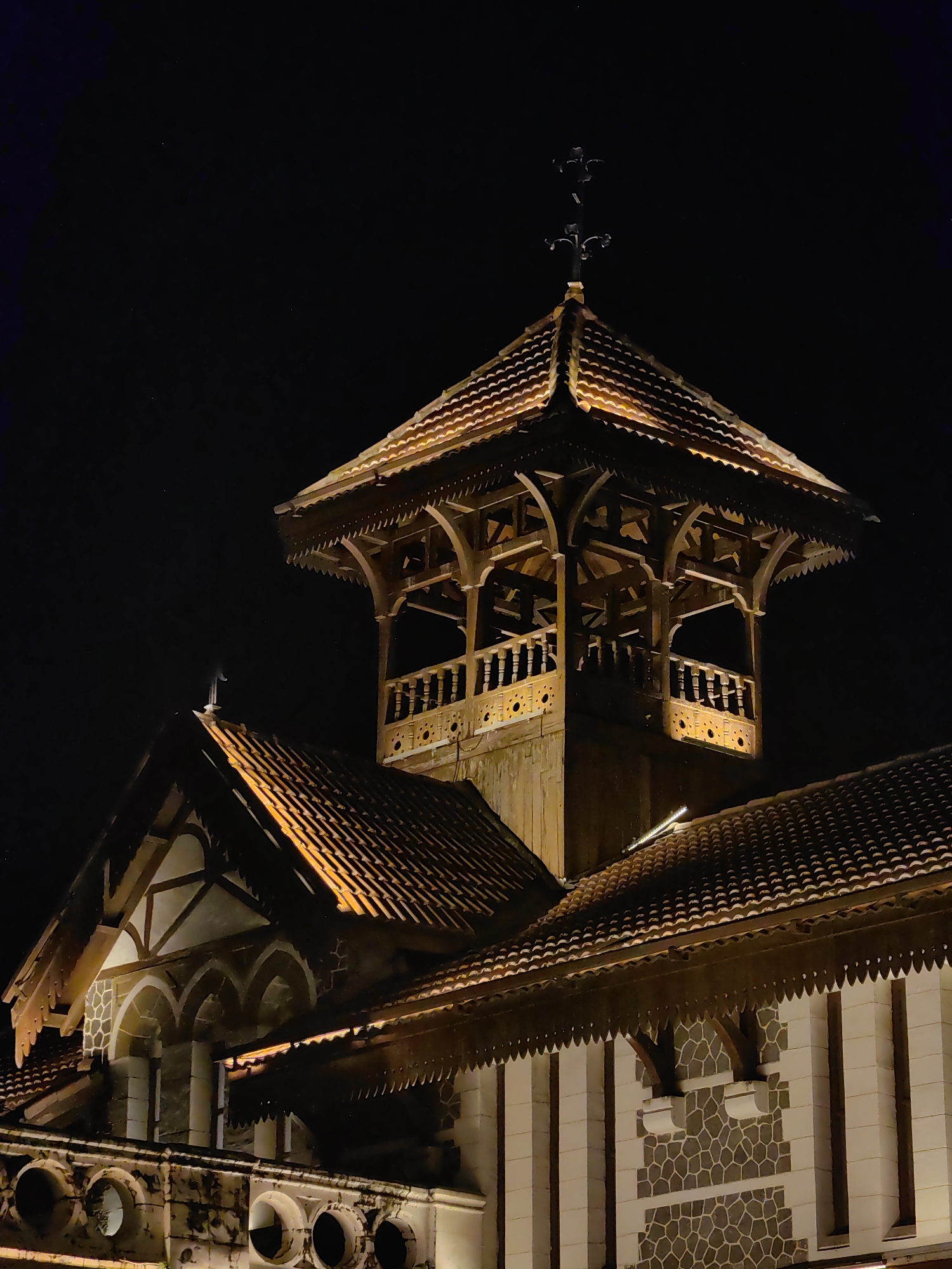 A nighttime, low-angle, medium shot captures the illuminated exterior of a historical building with a prominent bell tower in Mumbai, India. The building features multiple tiled roofs, some with ornate wooden eaves. The bell tower, constructed of wood and intricately carved, rises above the main structure and is topped with a cross. The exterior walls show a pattern of grey, irregular polygons against a lighter background, with arched windows visible on a lower section. The scene is dark, with the building illuminated by warm, artificial light.
